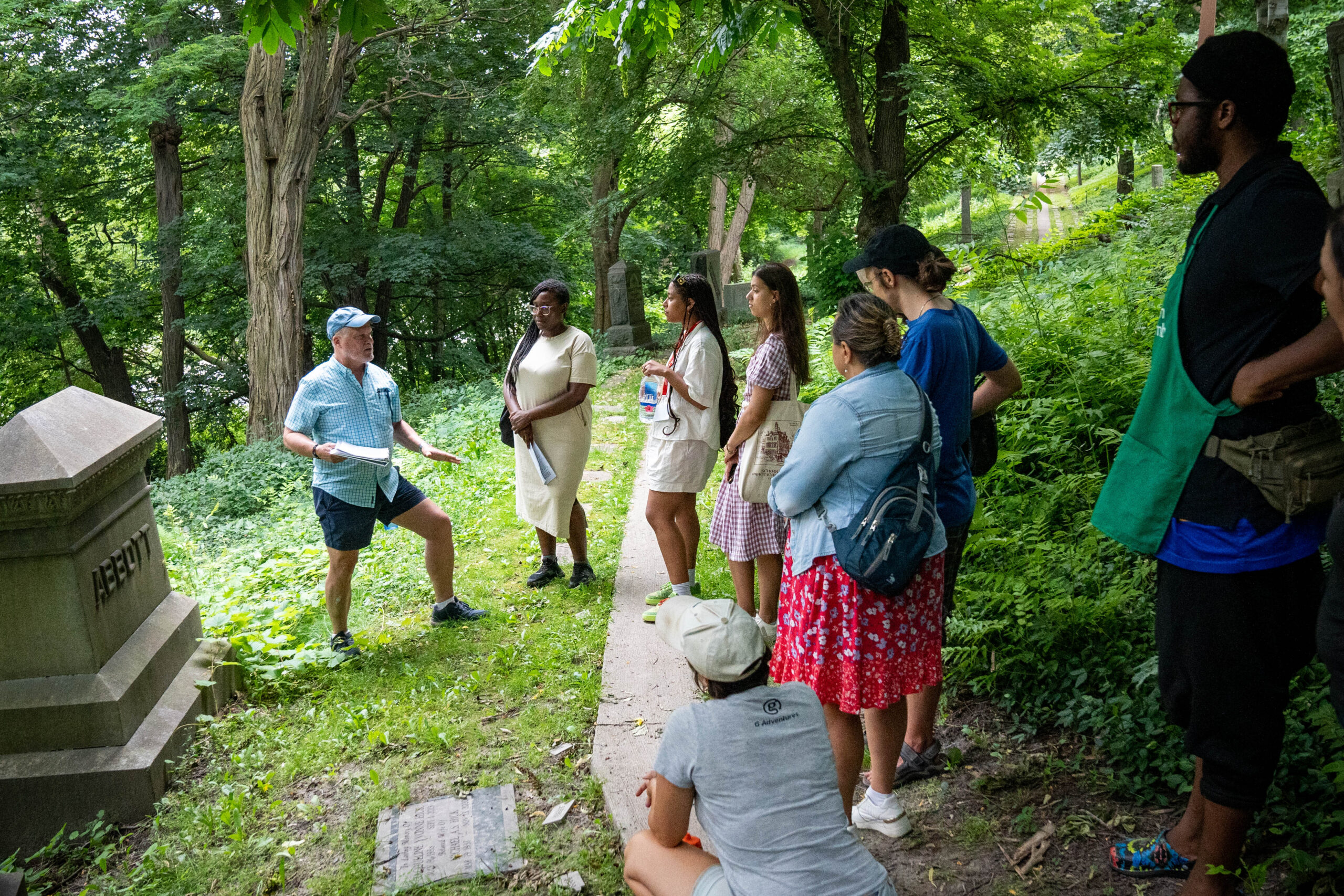 A group of 8 people stand facing a tombstone on which is carved ABBOTT.