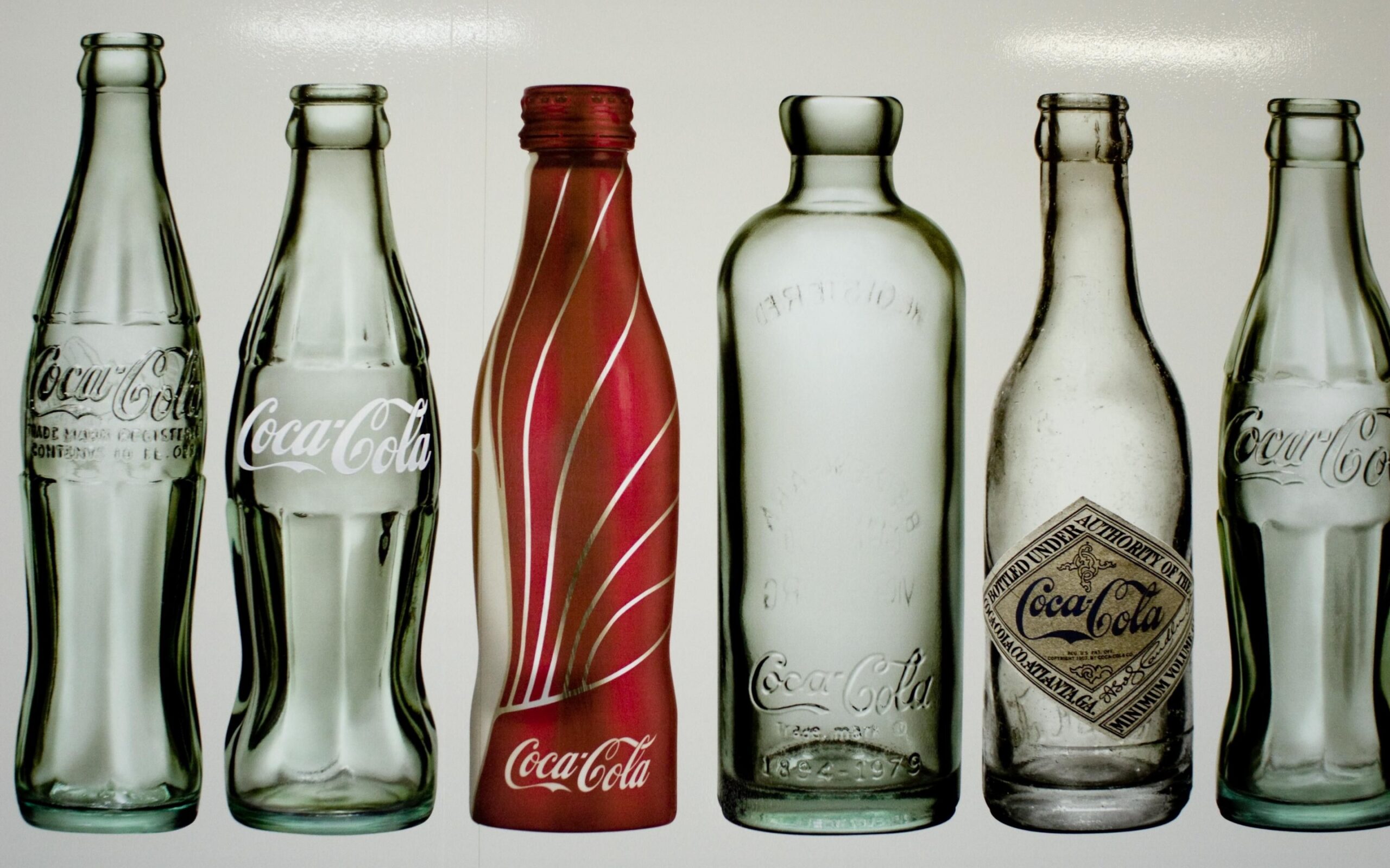 Six glass Coca-Cola bottles from unspecified sources. They are all approximately the same height. Three are similar to the modern glass bottle, while the other half are less familiar. One has a long neck and a slim but straight body. The next has straight sides and a very short neck. The final bottle is similar in shape to the modern glass bottle, but is mostly red with clear diagonal stripes.