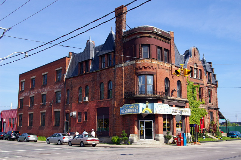 An old, red brick building on the corner of an intersection. There is a neon sign stating “Canary Restaurant.” There are a few cars parked next to the building, along with some people walking along the street.