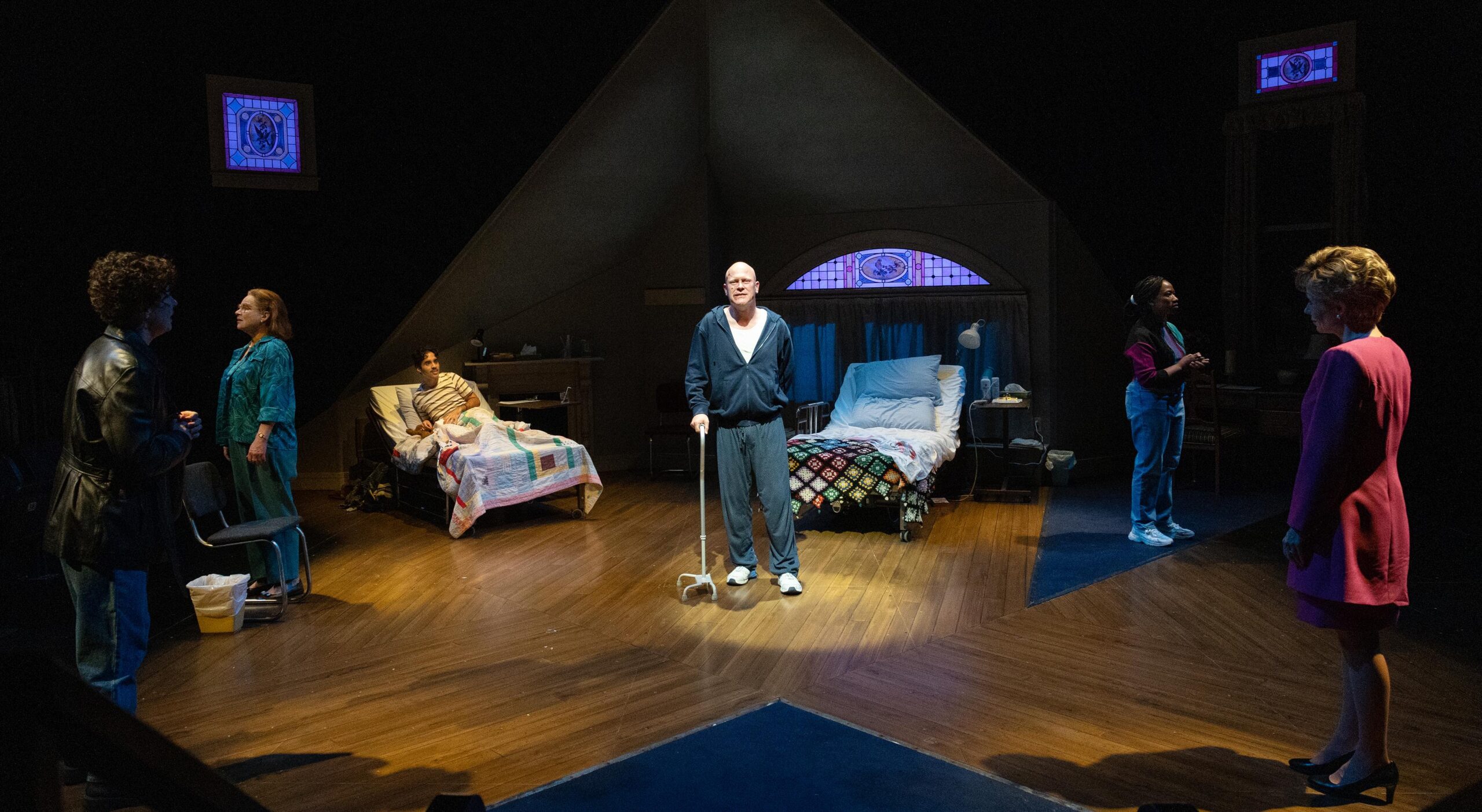 A play with a man standing in the centre with a cane and the spotlight on him, two women are speaking in the left corner, a person is laying in a bed in the back, and a woman in a pink blazer and skirt is staring at the man in the centre. His bed is behind him.