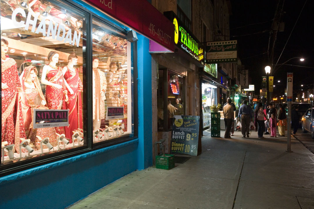 Photo faces down the sidewalk of Gerrard St E. showing the shop fronts. There is a shop with traditional South-Asian clothing in the display window with the name "Chandan Fashion". In the background a group of people crowd around the entrance to a restaurant.
