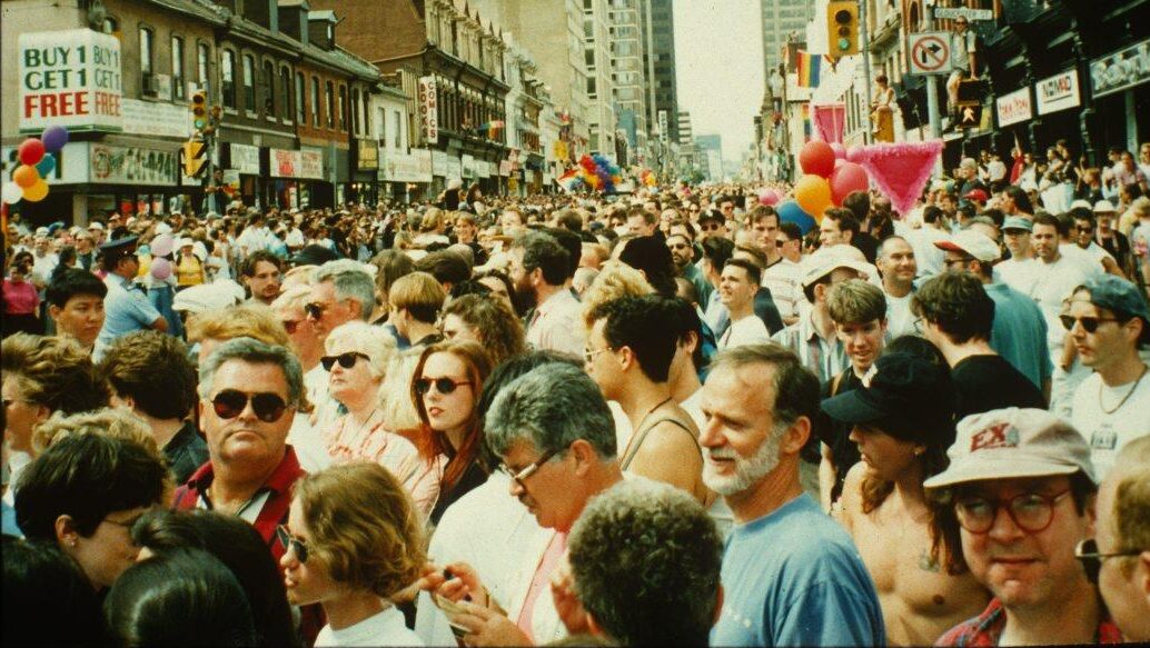 A photograph of a crowd on a street. Colourful balloons and an inverted pink triangle are visible. Commercial storefronts line the street.