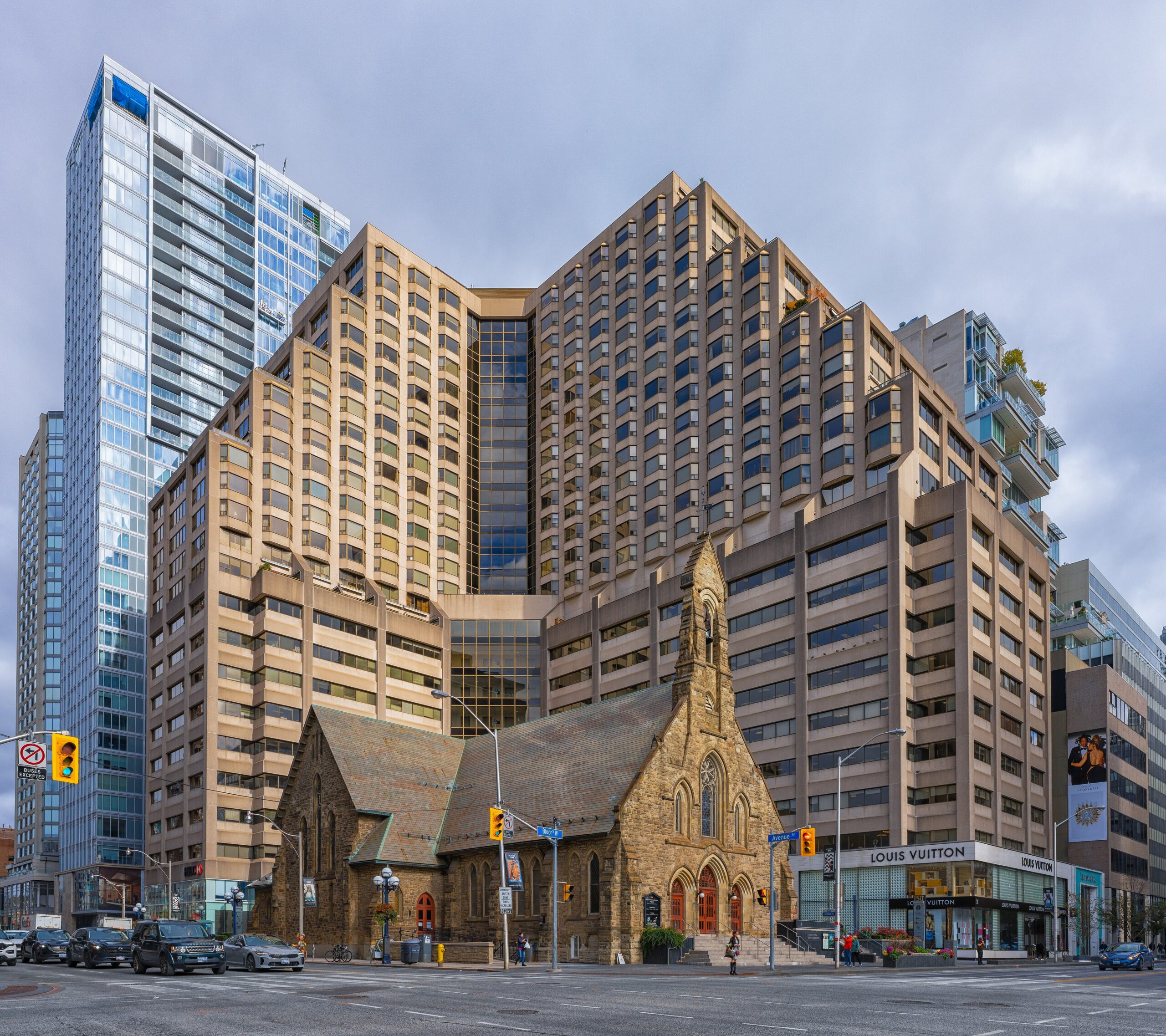 A Gothic Revival-style church building flanked by a large L-shaped high rise. More glass towers are seen the background.