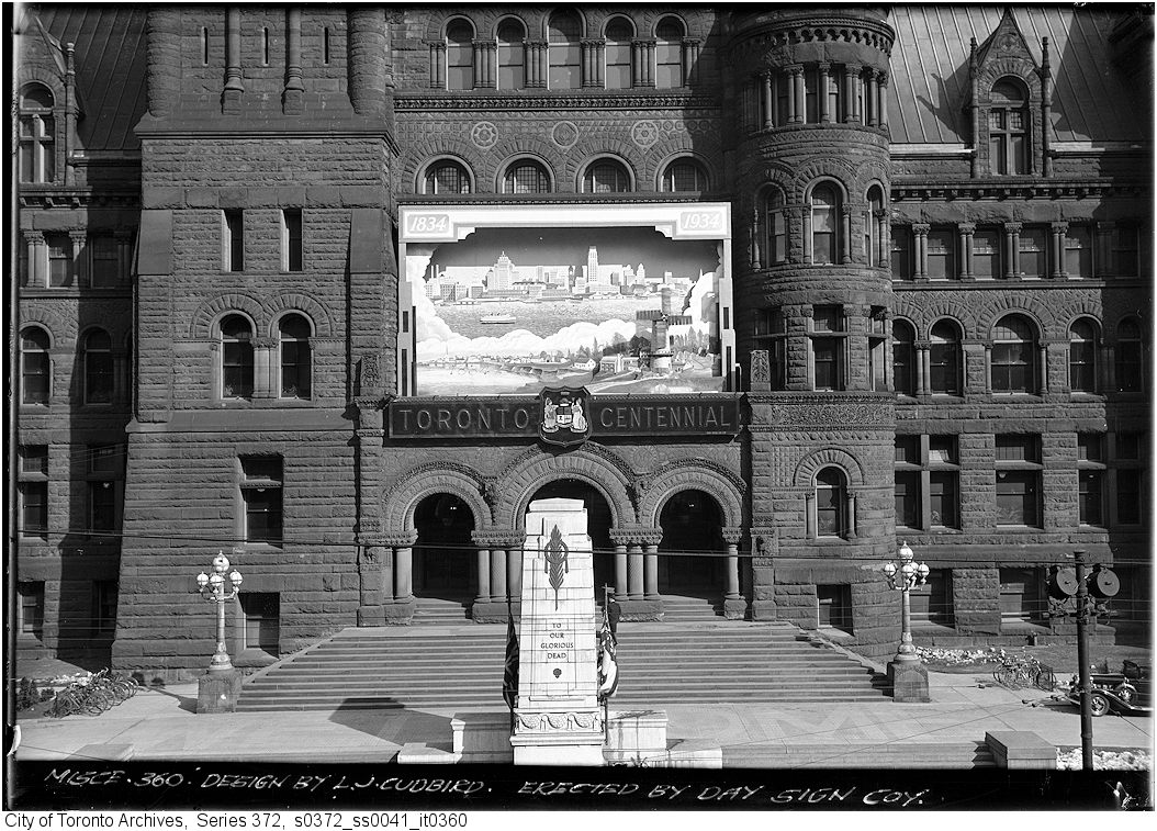 The façade of Old City Hall on a sunny day with the Cenotaph in the foreground. Affixed to the second and third floors above the entrance is a large sign depicting the city’s waterfront with the words Toronto Centennial and the city’s coat of arms beneath.