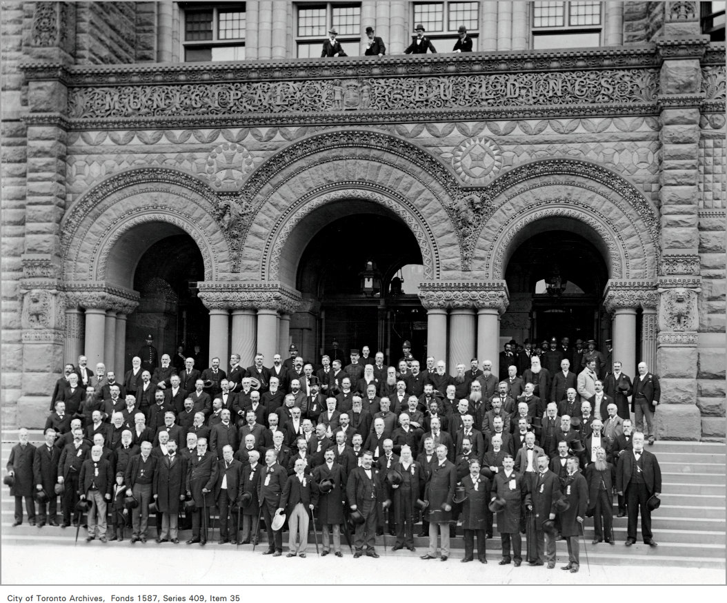 A group of about a hundred men dressed in Victorian formalwear stand on the steps of the main entrance of Old City Hall. The building’s ornate carved-stone archways are above them.