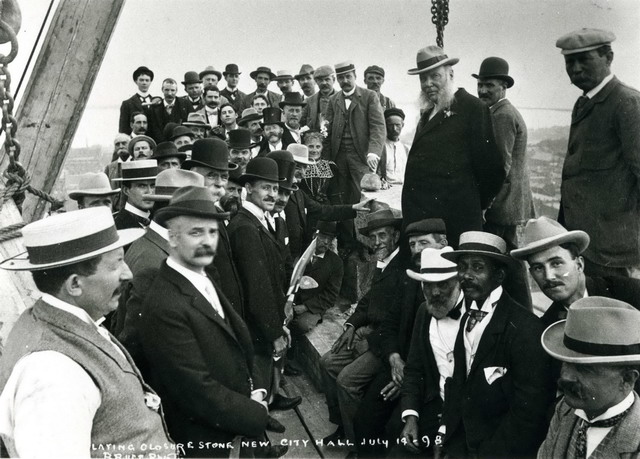 A group of men are gathered on narrow wooden walkway above a city skyline. Most are wearing hats and face the camera.
