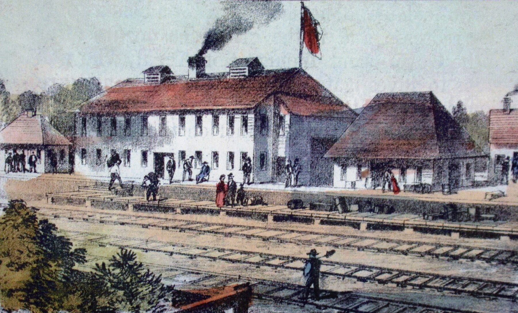 A colour illustration of the view of a two story building with smoke coming out of its chimney and a flag visible on the roof. There are other smaller buildings surrounding it, from the other side of the train tracks that line the complex.