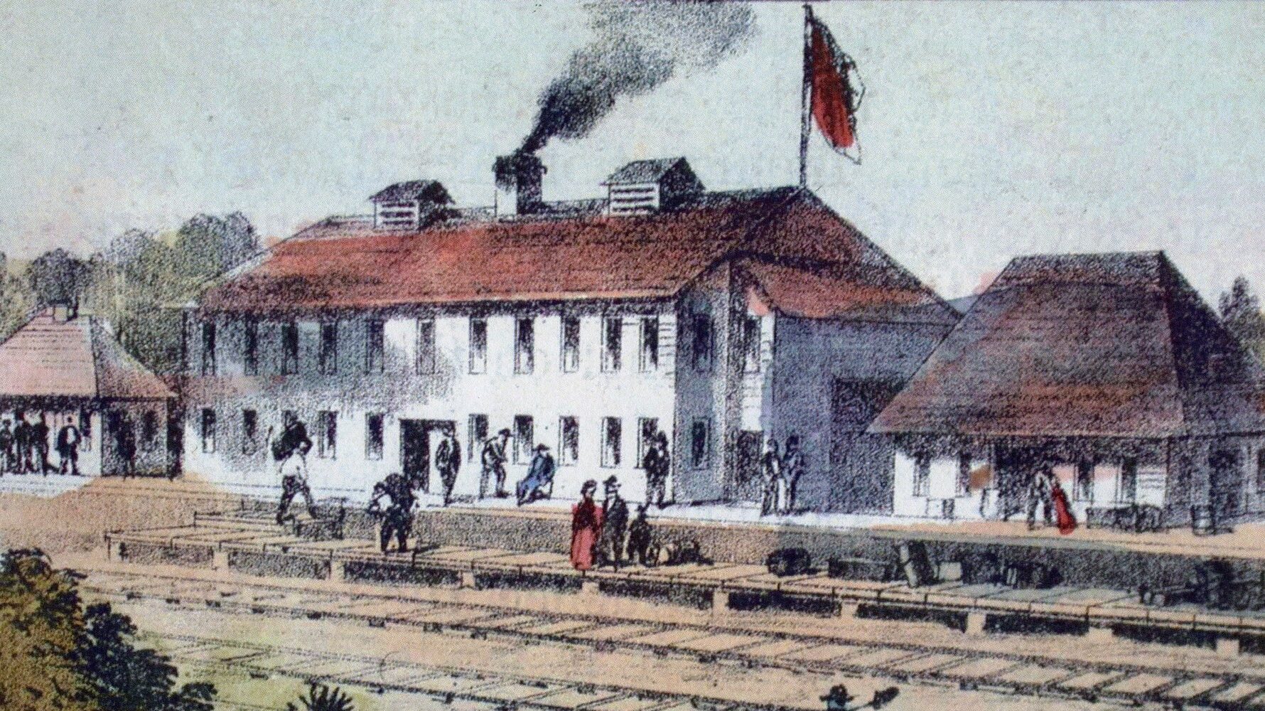 A colour illustration of the view of a two story building with smoke coming out of its chimney and a flag visible on the roof. There are other smaller buildings surrounding it, from the other side of the train tracks that line the complex.