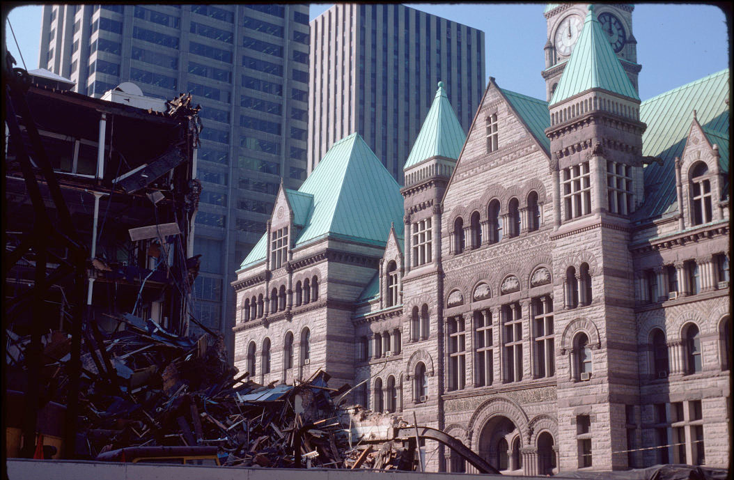 A view of the James St. façade of Old City Hall on a sunny day. High rises south of Queen St. are visible in the background. In the left foreground is a partially demolished building and debris.