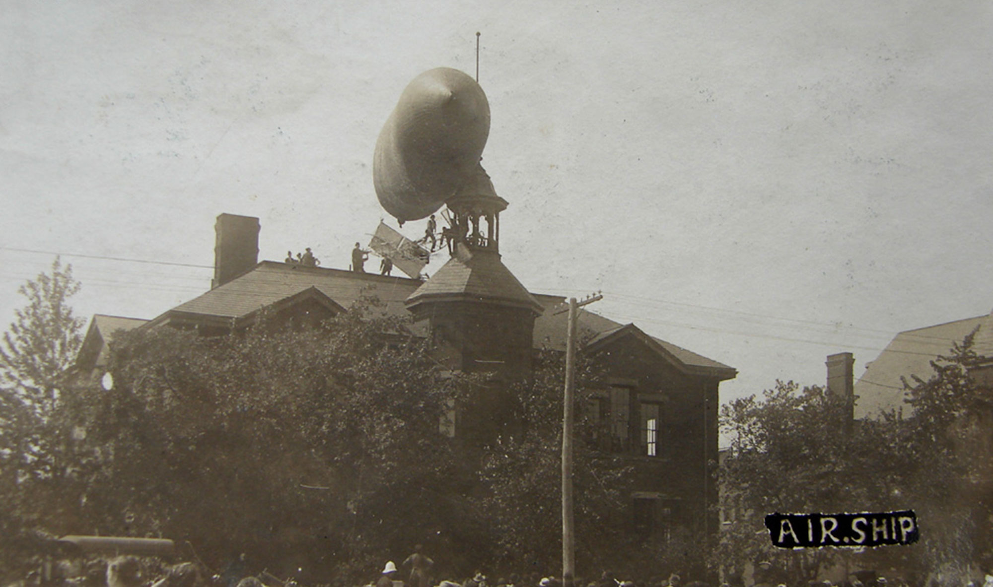 Black and white image of a small airship tangled on the roof of a building with a crowd gathered below