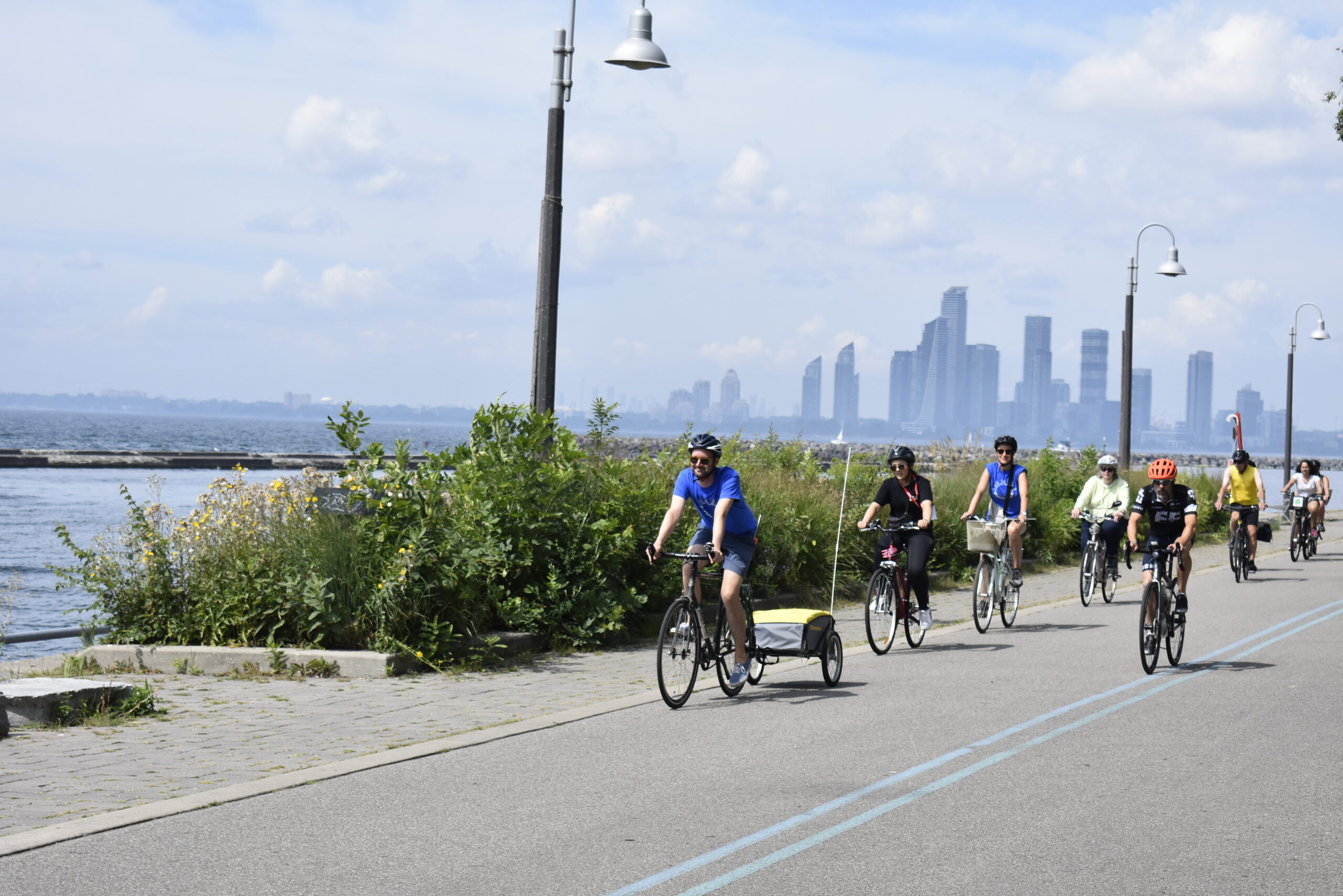 Line of cyclists riding on the side of a round next to a waterfront. In the distance a cityscape is visible of tall skyscrapers.