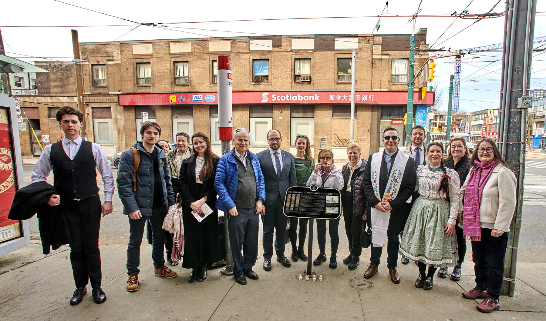 Image of 14 people, some in traditional Hungarian costume, standing on a sidewalk beside a historical plaque.