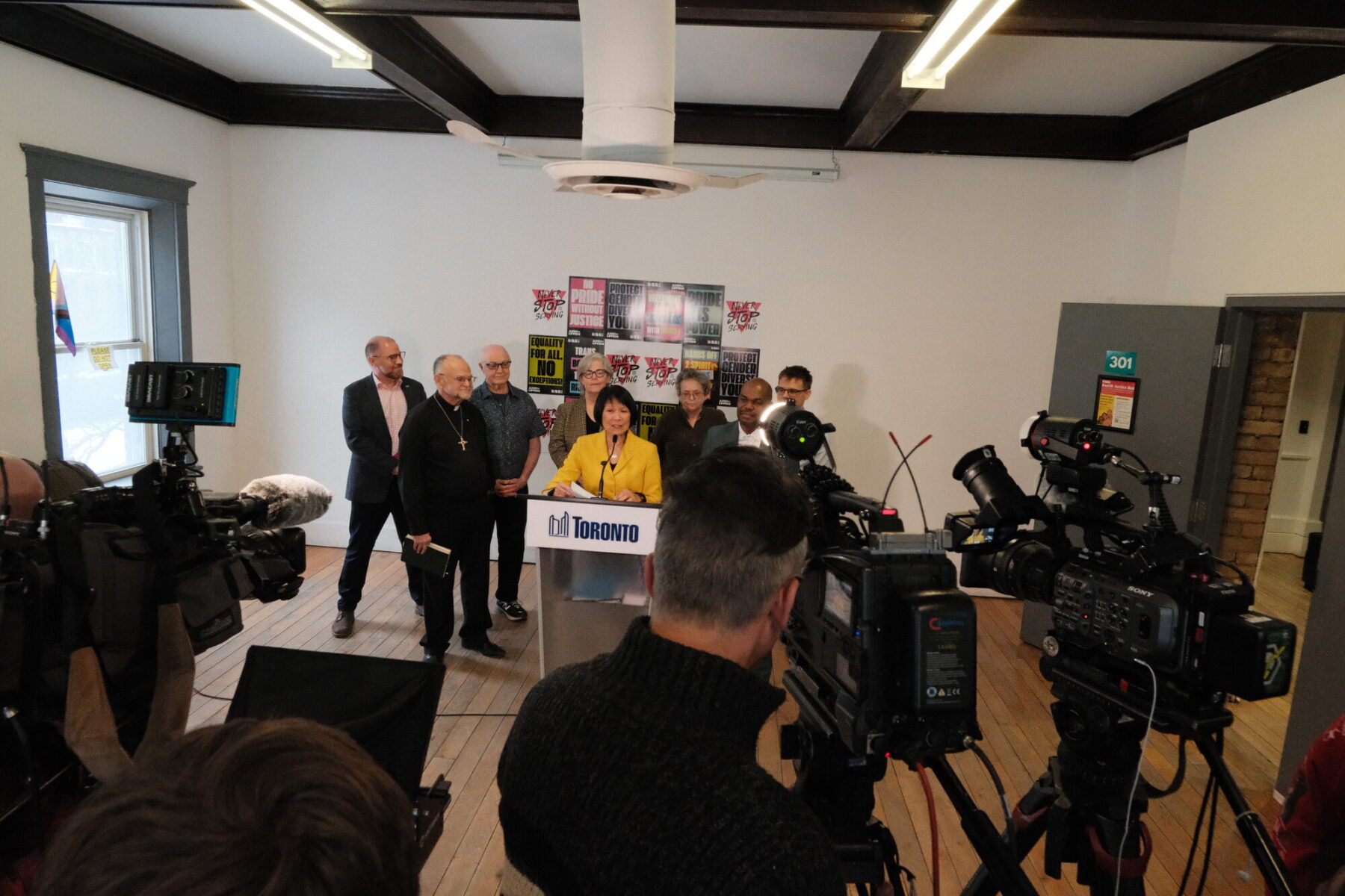 Woman speaks into a microphone at a podium with the City of Toronto logo. Behind her seven people are standing. In front we see videographers and cameras.