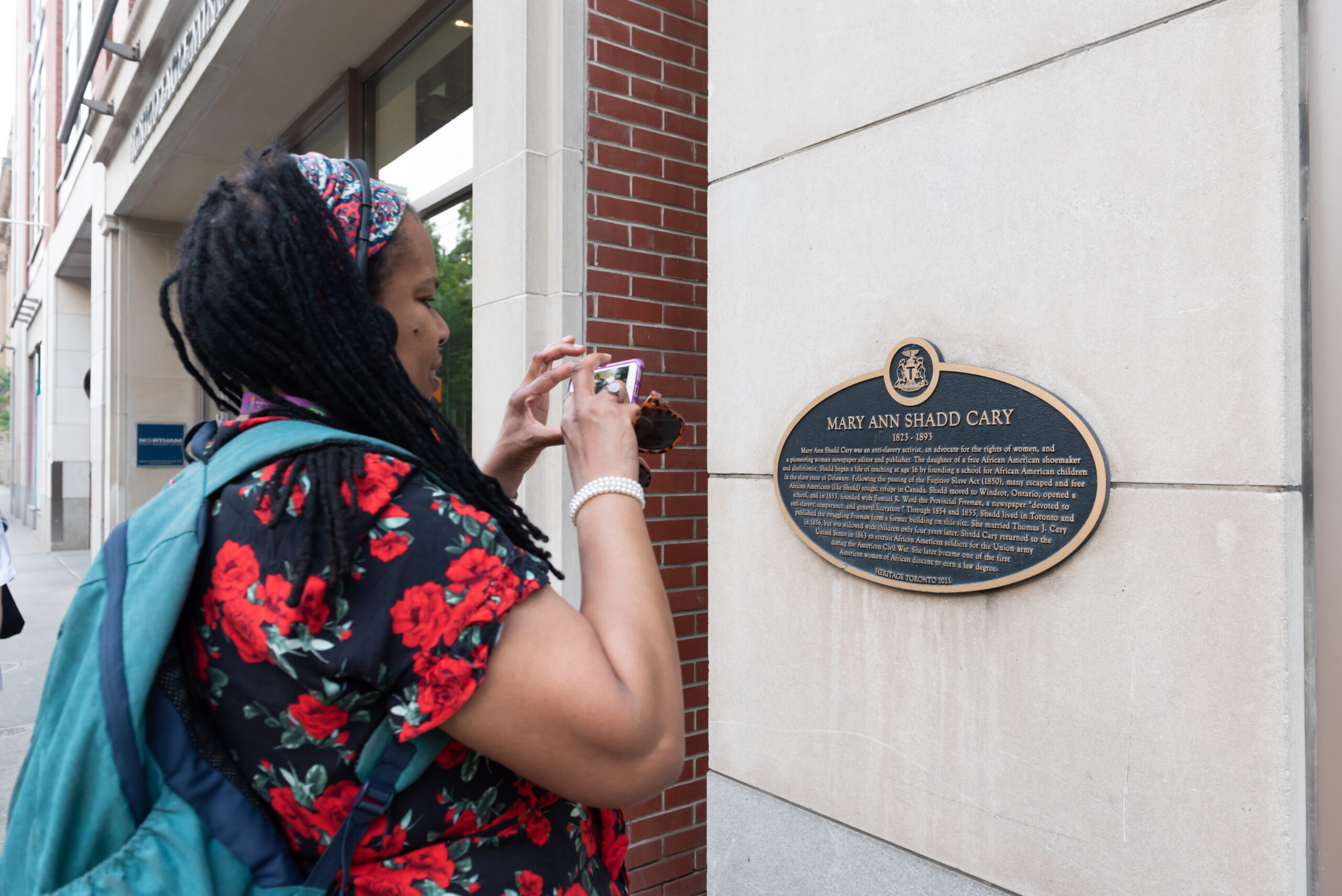 A woman holding a digital camera takes a photograph of a plaque on a wall.
