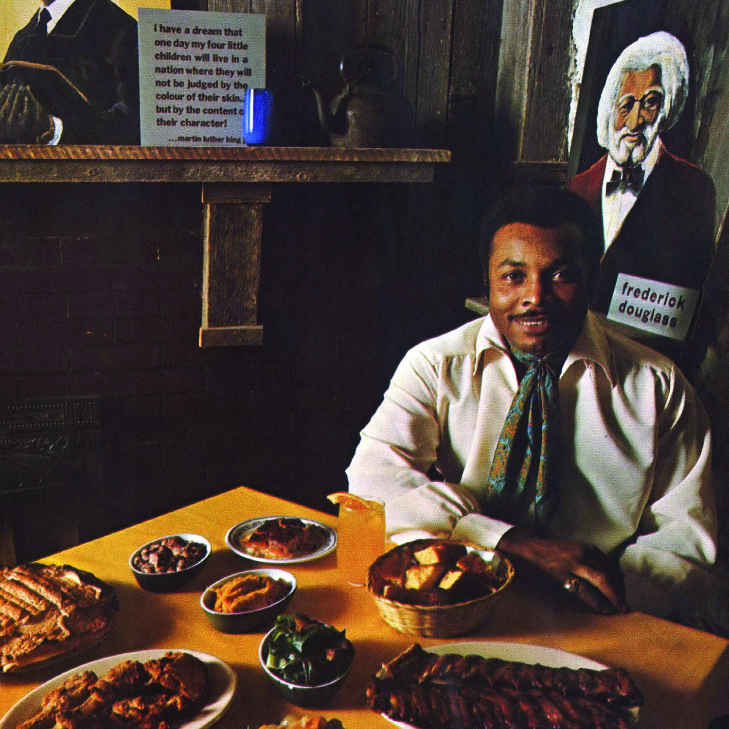 Image of a man seated at a restaurant table with items of food laid out in front of him. Paintings of Frederick Douglass and Martin Luther King, Jr. are on the wall behind him.