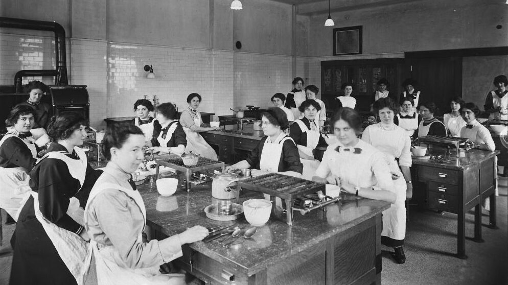 Twenty two young women sitting at large tables in a domestic science class. They all wear white aprons and have their hair tied back. On the tables are gas cookers and various utensils, bowls, and pots.