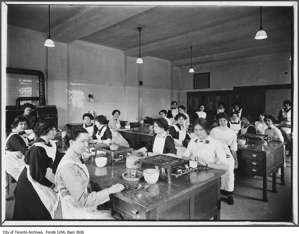 Twenty two young women sitting at large tables in a domestic science class. They all wear white aprons and have their hair tied back. On the tables are gas cookers and various utensils, bowls, and pots.