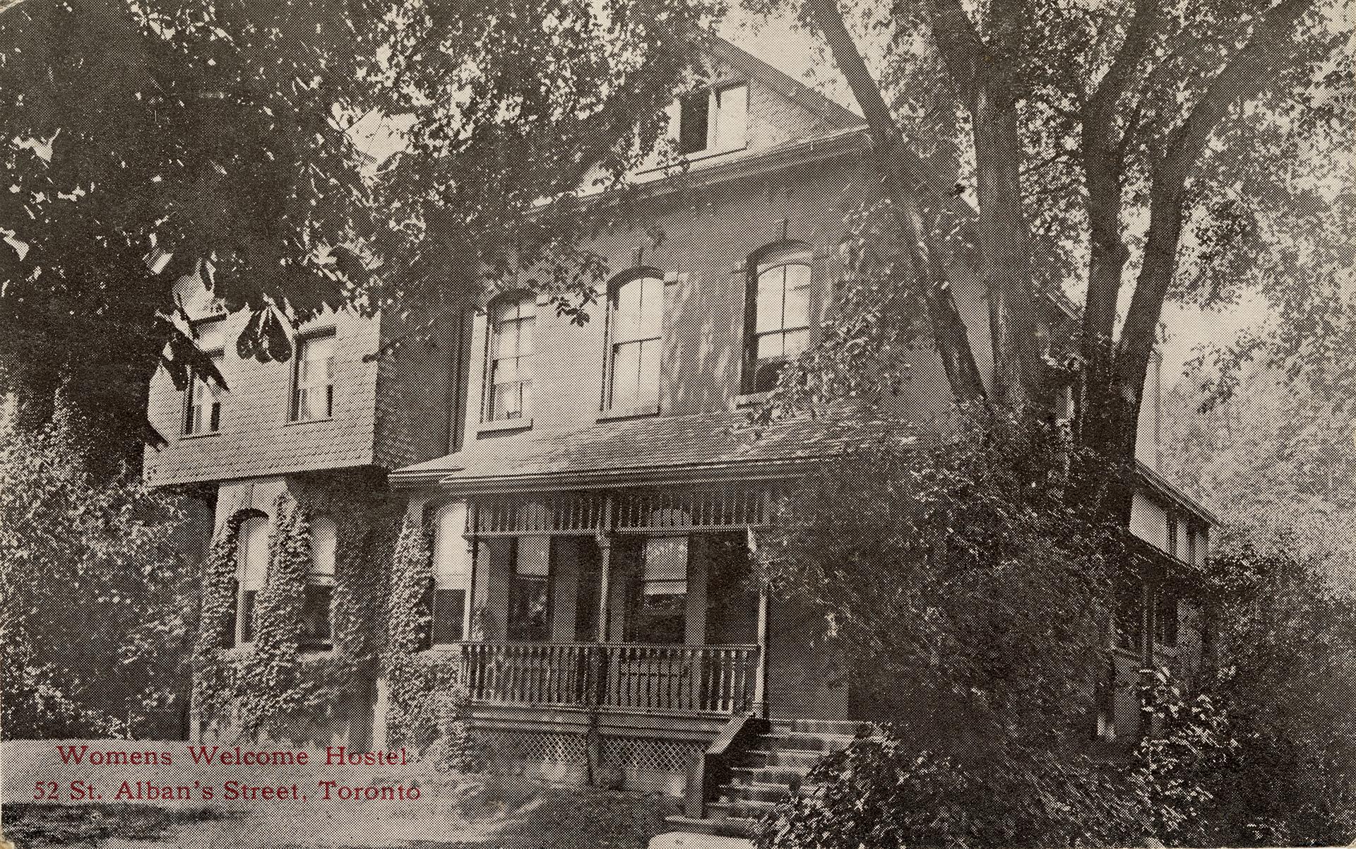 A large Edwardian house with stairs leading to a wide front porch. The far side and neighbouring house are partially covered with ivy. A large tree stands before the stairs. Red text in the lower left corner reads: “Womens Welcome Hostel, 52 St. Alban's Street, Toronto."