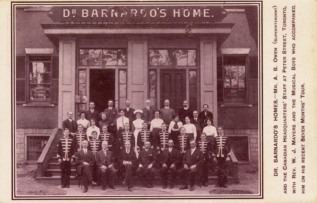 A group of staff and a visiting musical group stand outside a building. A sign on the building reads, “Dr. Barnardo’s Home.”