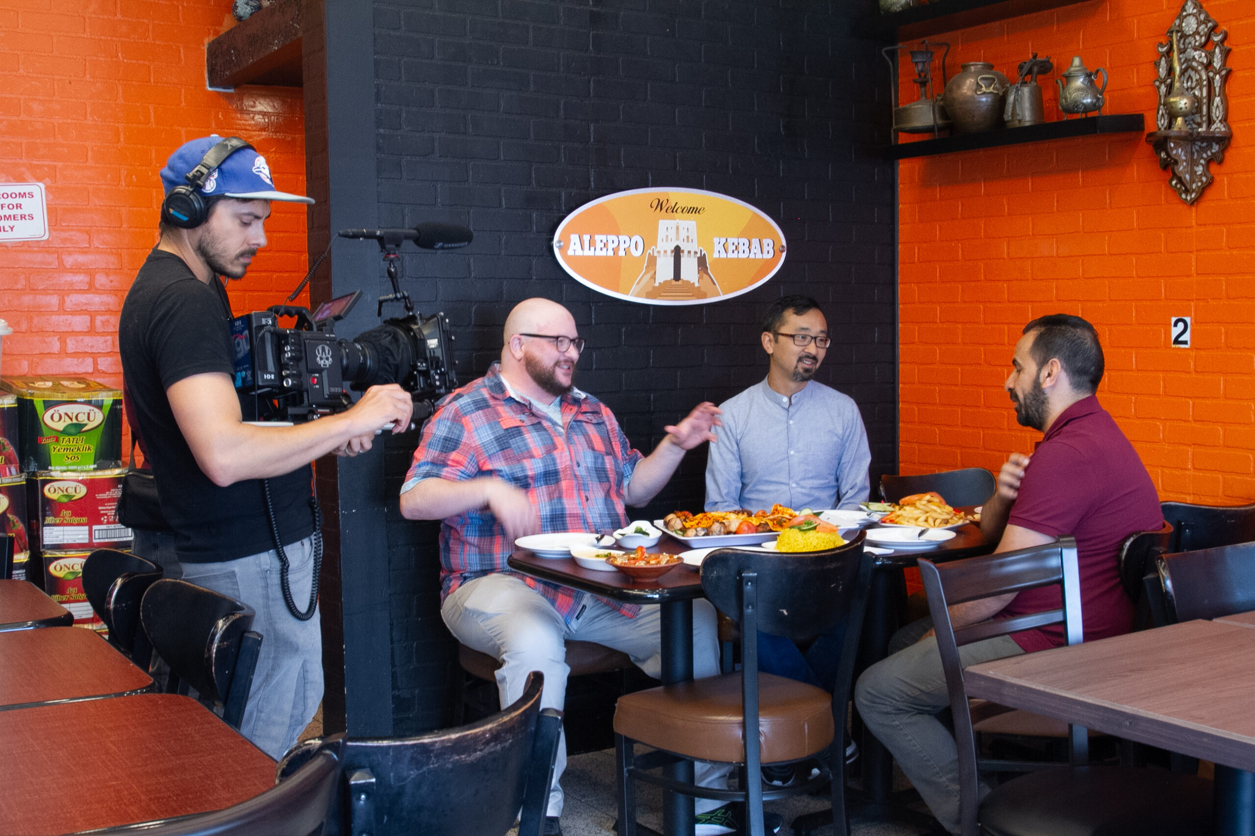 A group of three men sit at a table together. A fourth man stands near the table with a large camera as he films them. The brick walls surrounding the table are painted black and orange, and a sign hung on the wall reads "Welcome Aleppo Kebab."