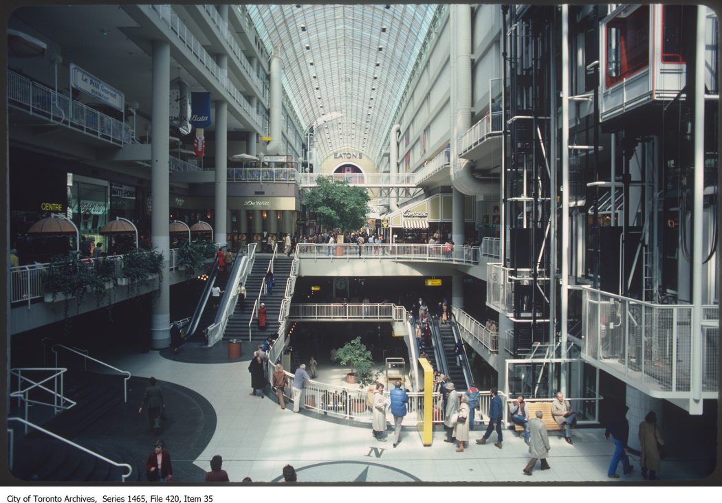 An interior view of a multi-storey mall. The ceiling is rounded and made of transparent glass, allowing light to shine into the building. Elevators and escalators can be seen moving shoppers between the mall's storeys.