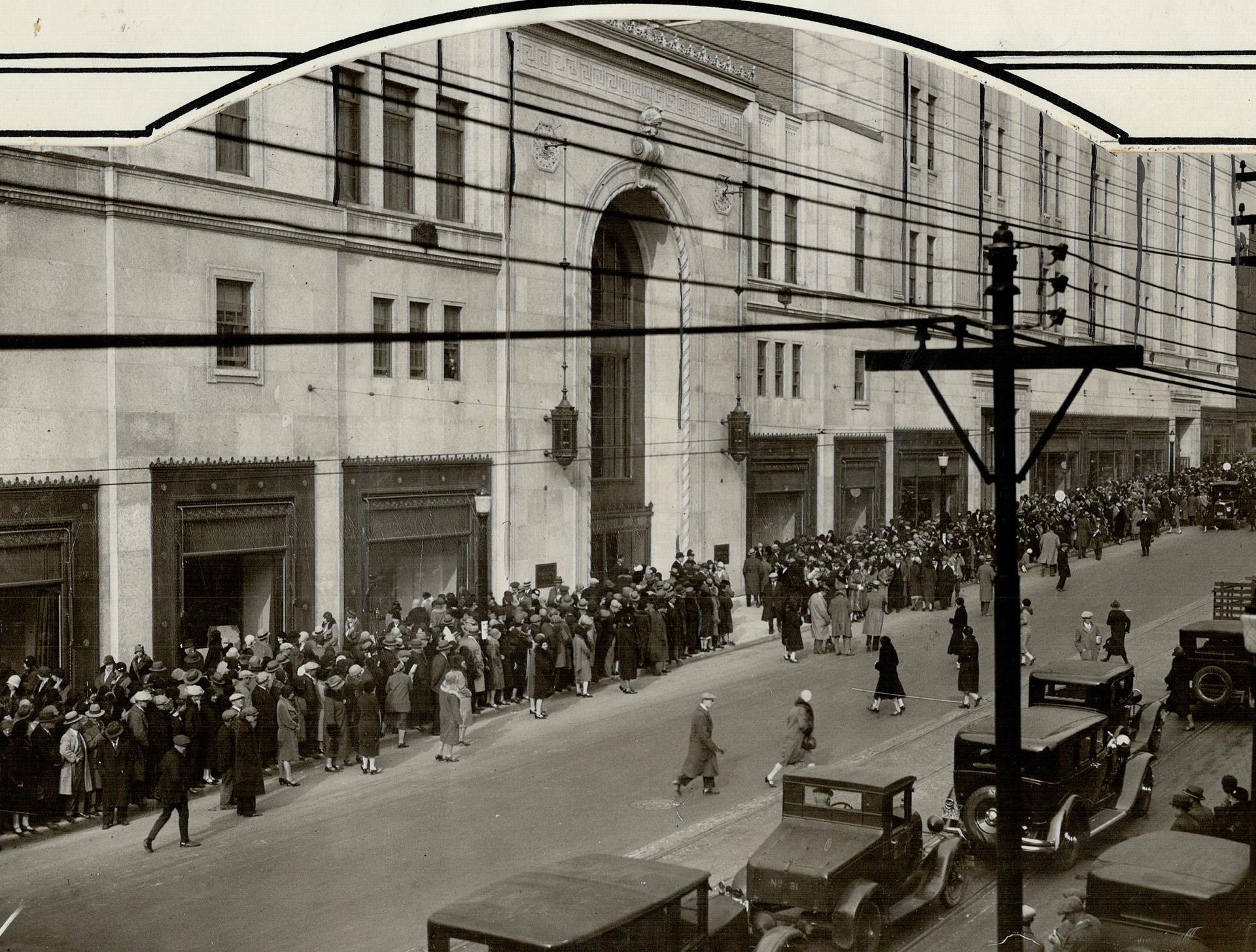 A large crowd lines a busy urban sidewalk on both sides of a grand arched entryway. Shop windows of a department store can be seen behind the crowds. In the foreground are historic cars.