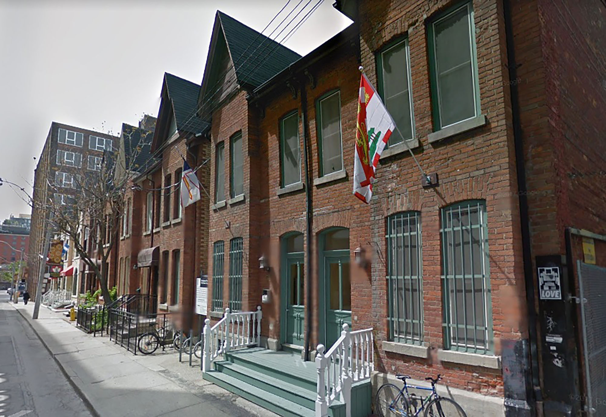 A street scene showing several red-brick row homes. Flags are hung outside the first two homes closest to the camera. Three steps can be seen leading to the front doors of the homes.