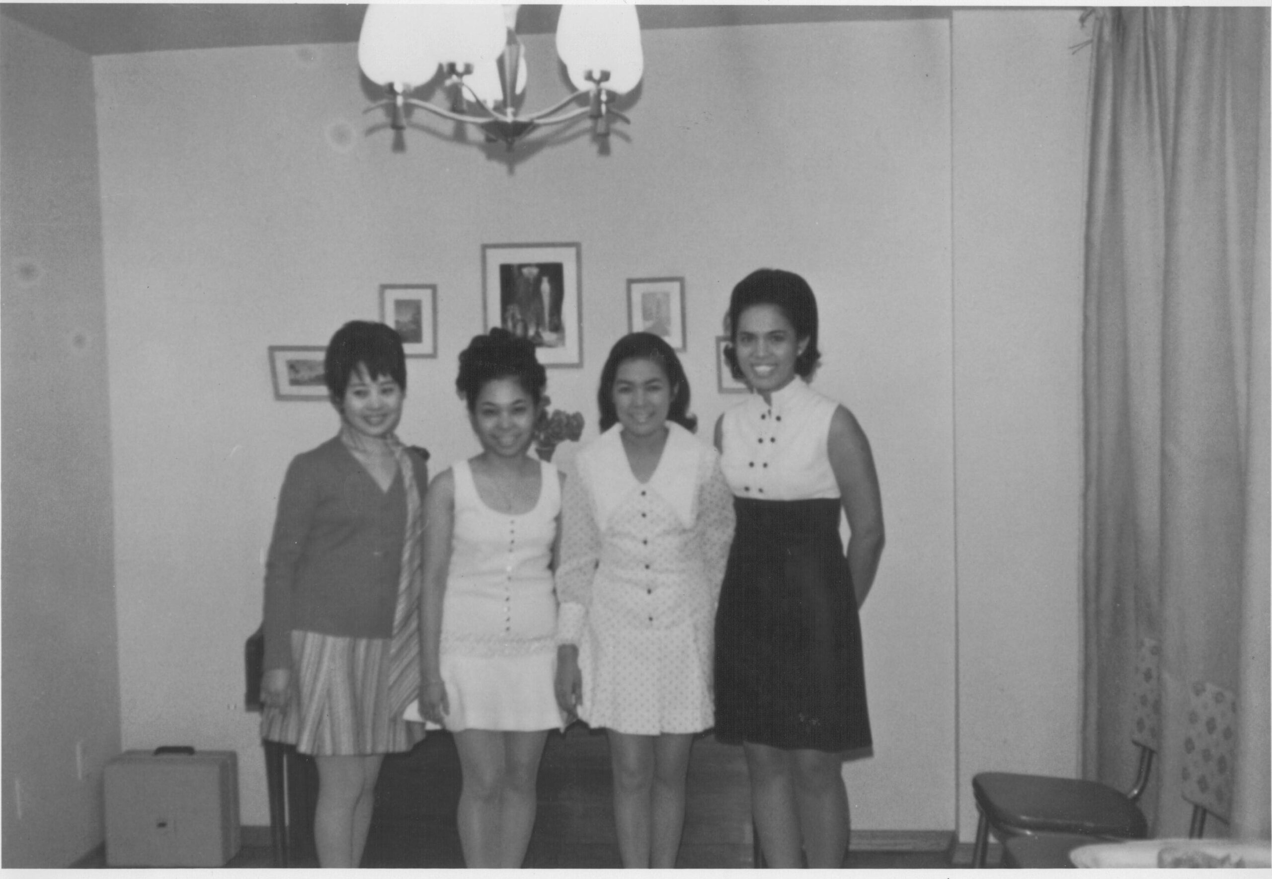 A black and white image of a group of four Filipino women standing together smiling and posing for the photograph in what looks to be a living room.