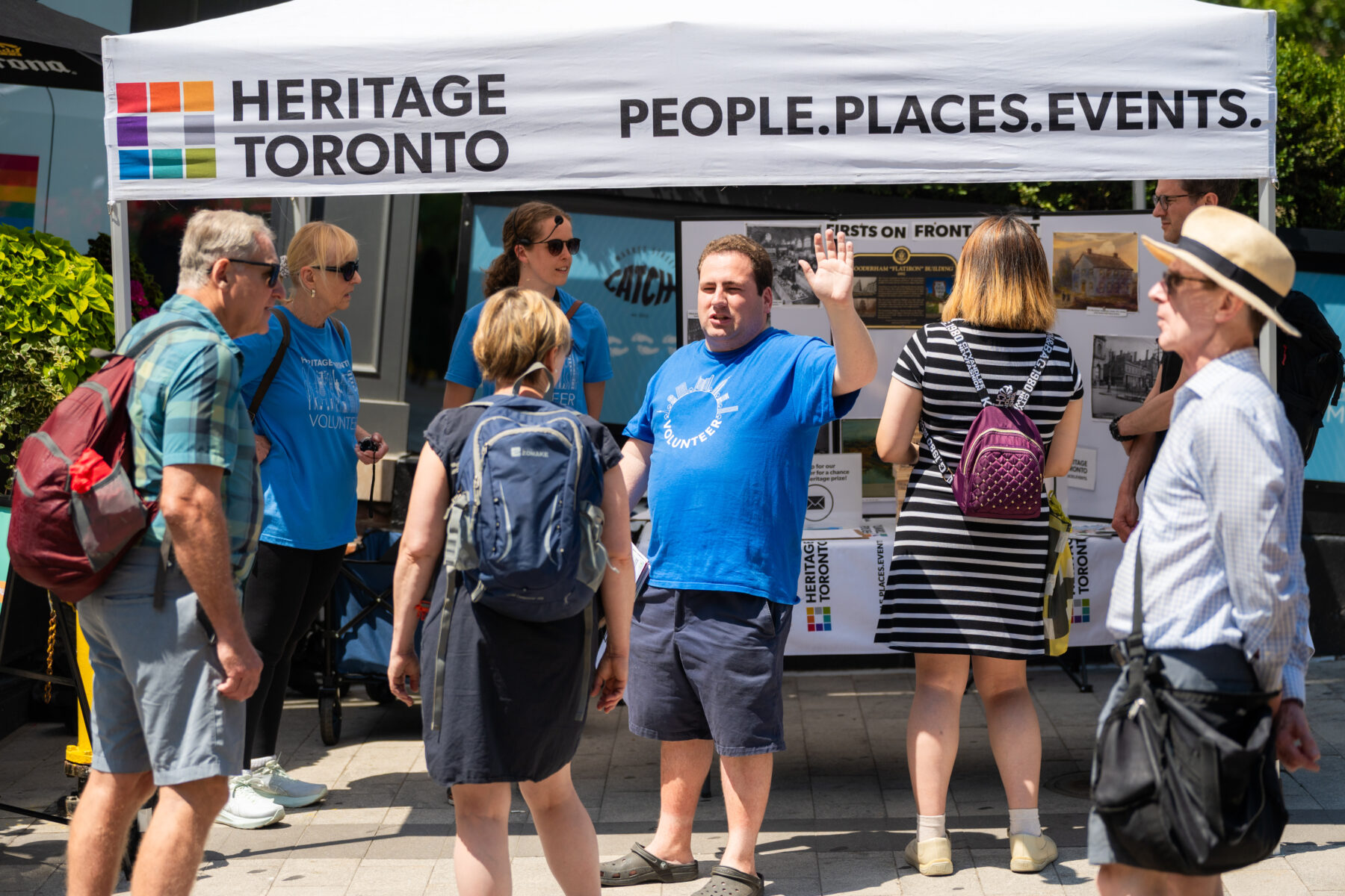 Next to and under a tent with the Heritage Toronto logo, eight people are gathered. The one in the middle faces the camera, wears a blue tshirt, and has his right hand raised.
