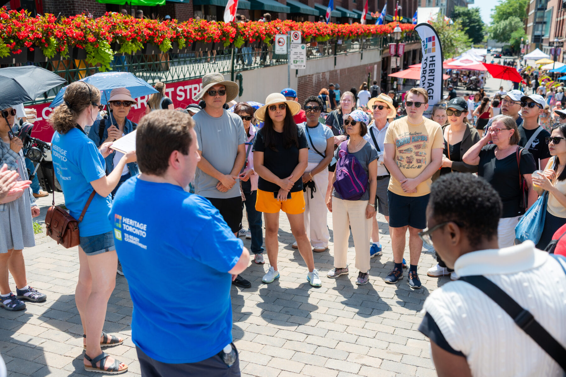 Group of people stand on cobblestones . Behind them a long railing full of container plants is visible. In the background tent canopies can be seen.