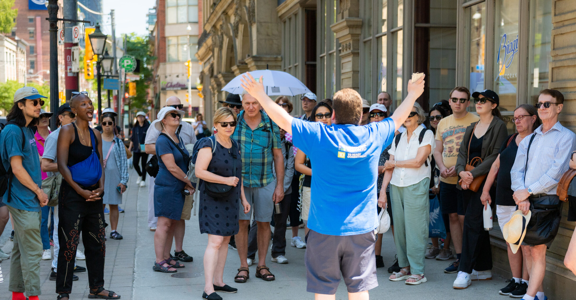 People in summer clothing stand on a sidewalk. A person in the front with the back to the camera raises both hands in the air. To the right, an ornate stone and glass faced building appears.
