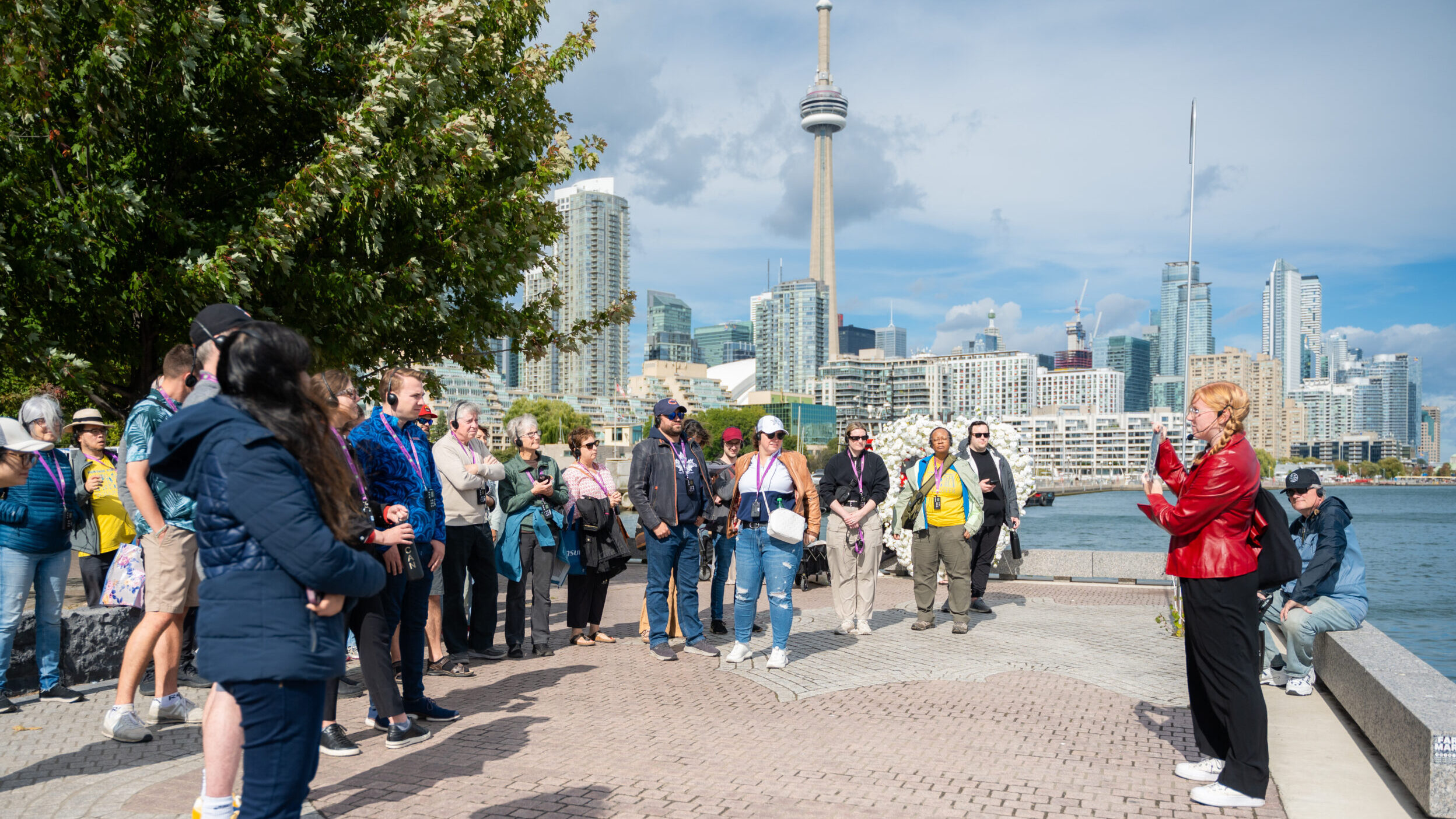 A large group of people stand on a pedestrian walkway listening to a speaker on the right. In the background, a body of water is visible along with a cityscape showing tall buildings and the CN Tower.