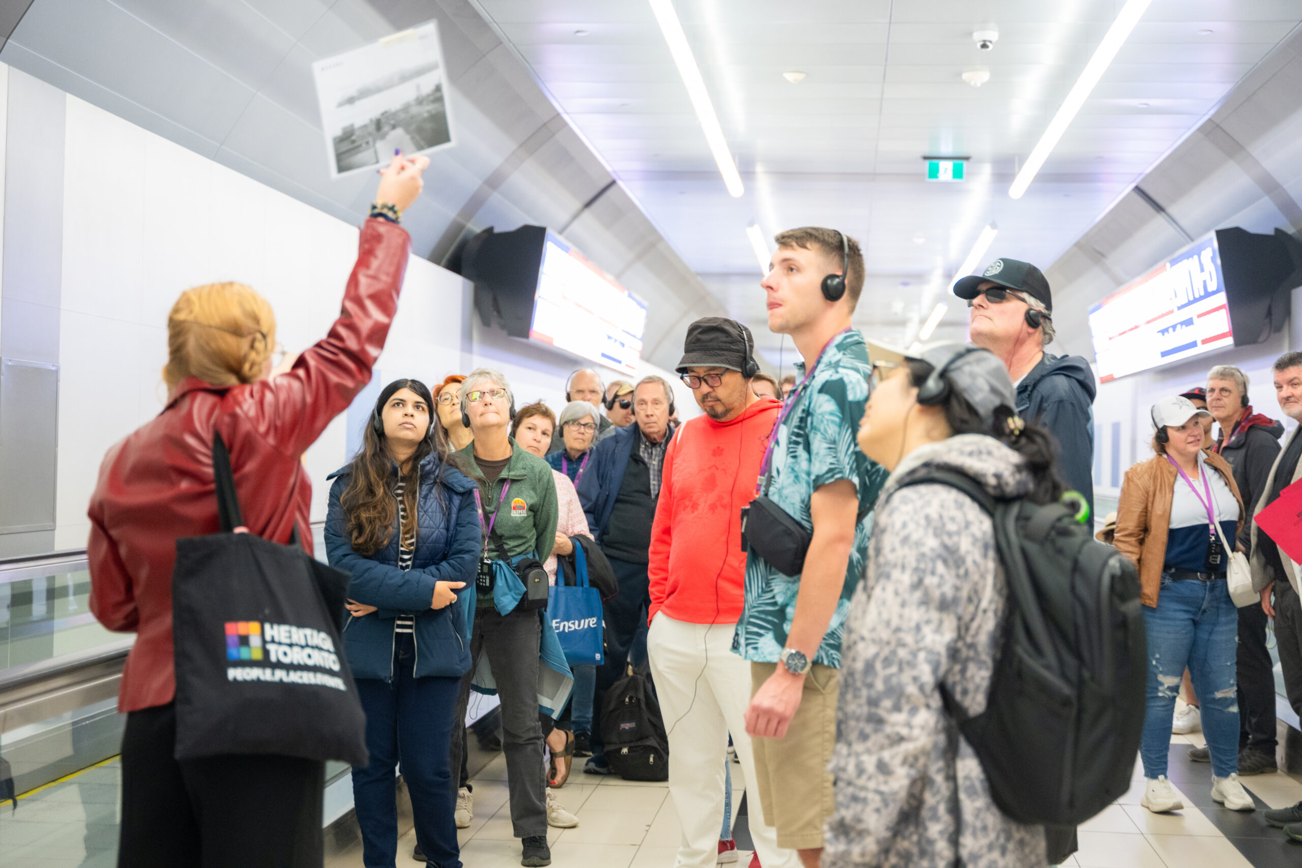 A group of people look up at a small poster held up by a guide. They are inside an airport corridor.