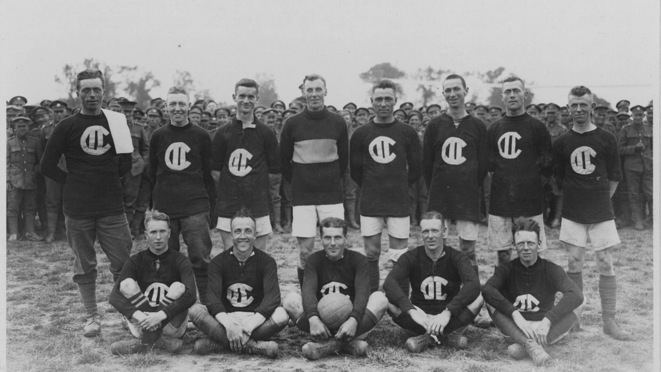 Two rows of 13 people wearing sports uniforms posing for a picture. One person in the centre is holding a soccer ball.