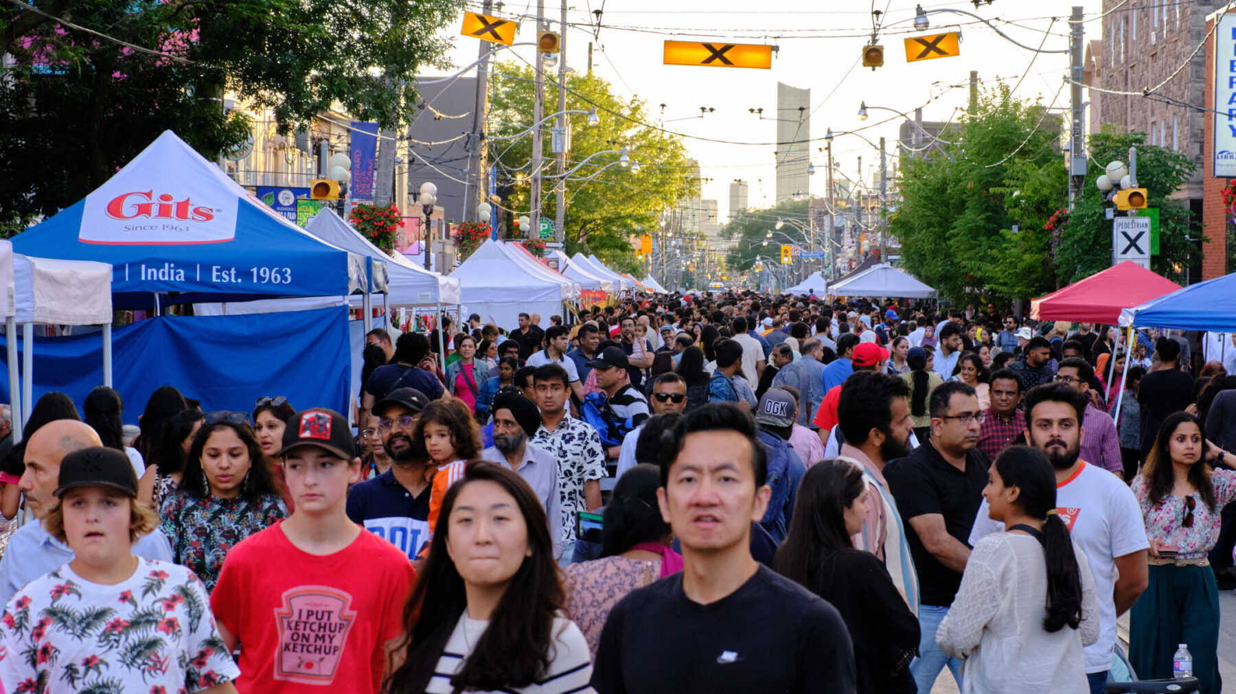 Photo faces west down Gerrard St E. at the intersection of Gerrard St and Ashdale Ave. Tented booths with various vendors line both sides of the street and the road is filled with a large crowd of people visiting the festival.