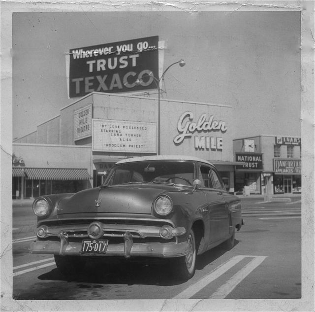 Black and white photo of an old-fashioned 1960s car parked in front of a shopping centre with many signs. The main sign reads, "Golden Mile". Other signs read, "Wherever you go...trust Texaco", and "National Trust".