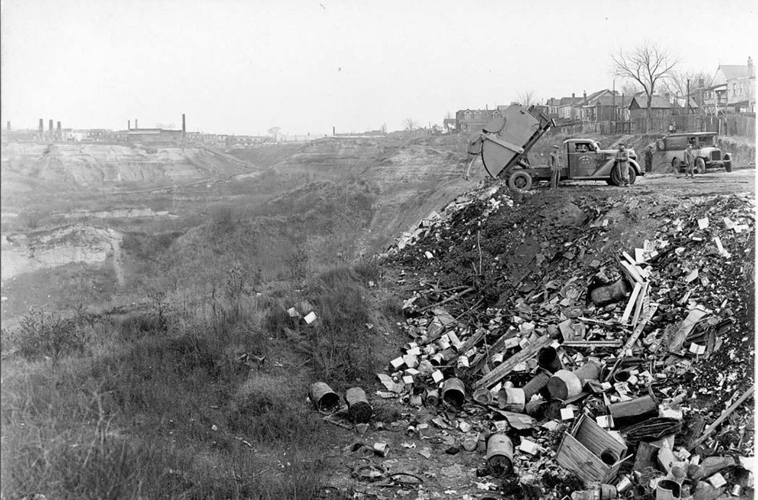 A black and white image of a dumpsite with trucks dumping waste (mainly bricks and buckets) into the pile of garbage.