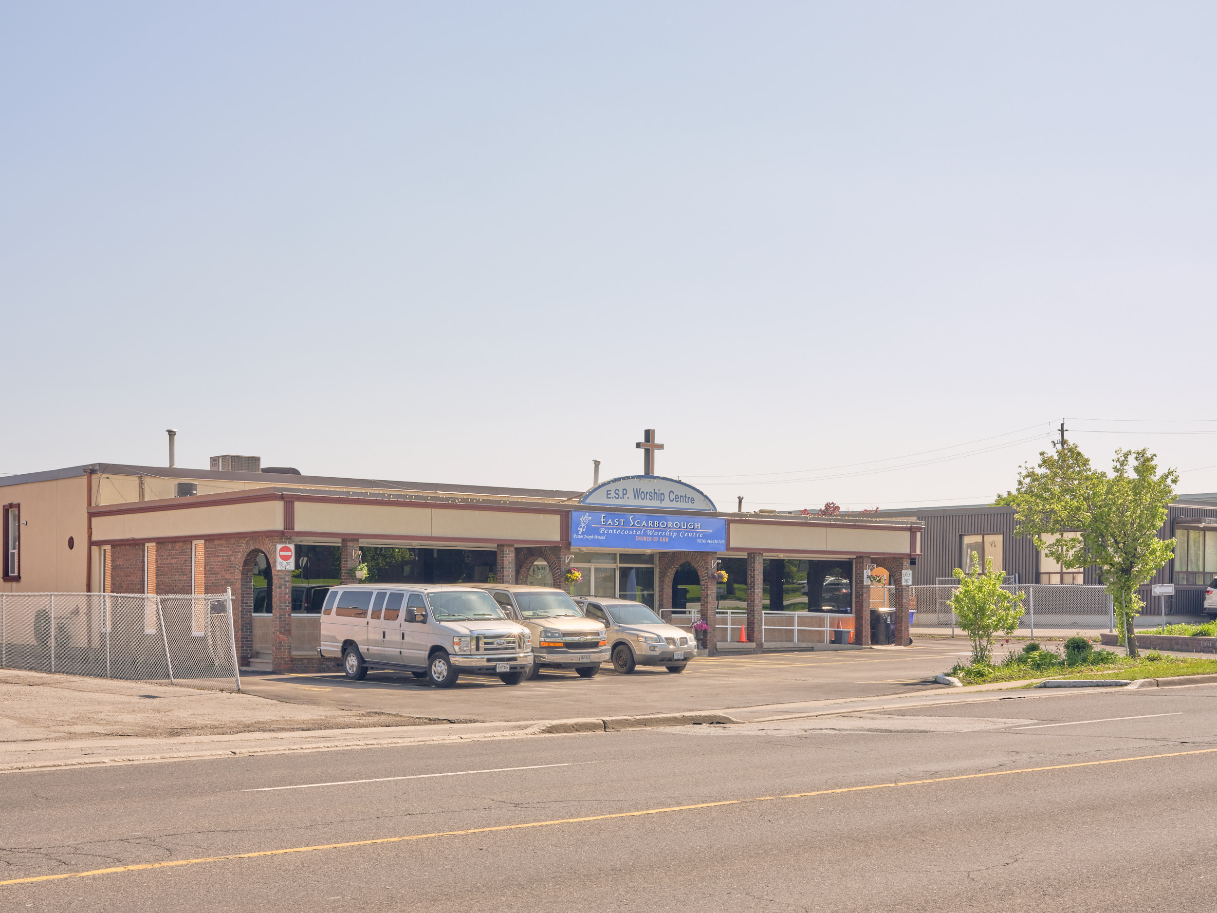 Street view image of single story building with a curved signboard and cross on the front façade. There are 3 white vans parked on the left in front of the building and small trees on the right side.