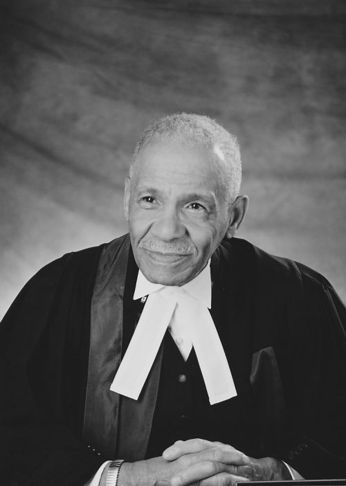 A man sits facing the camera wearing the formal robes of a judge. His hands are clasped in front of him.