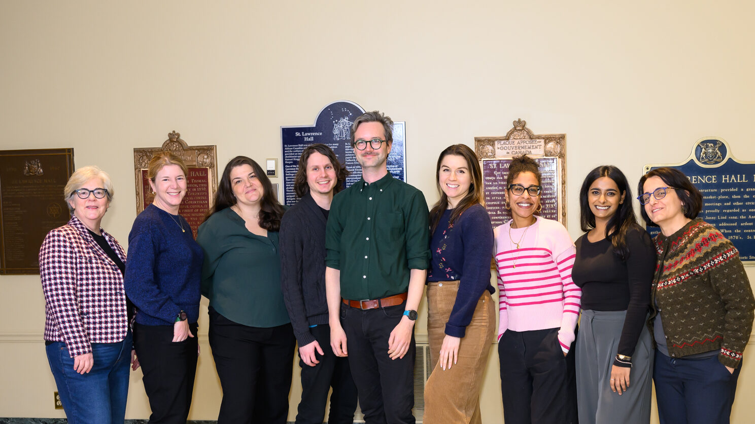 Photo of eight people smiling and facing the camera. Behind them several plaques are mounted on the wall.