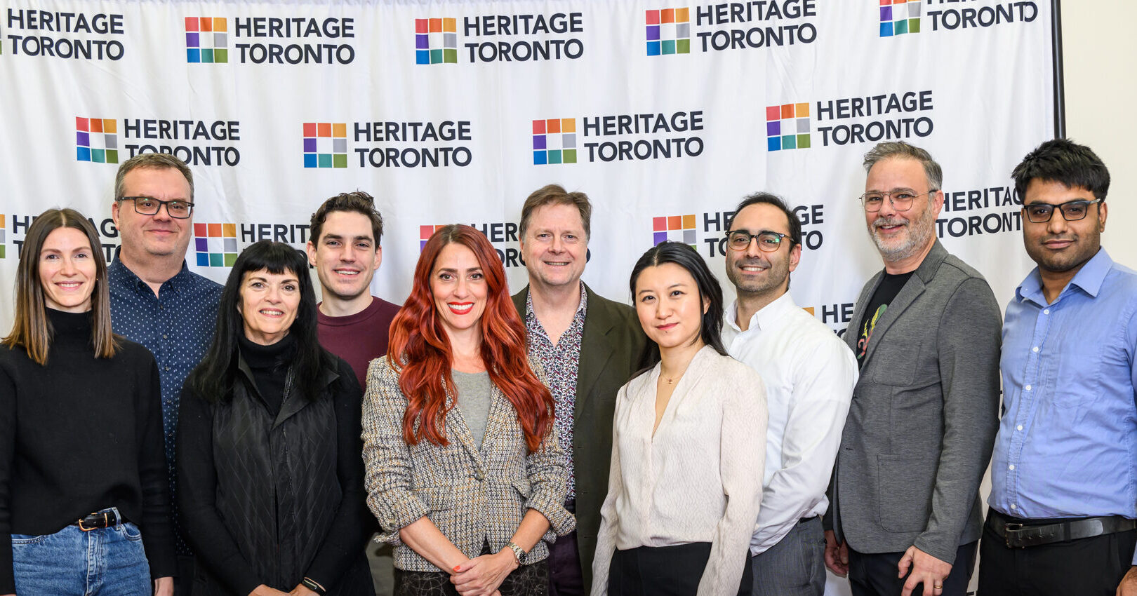 Group of people stand in two rows, smile and face the camera. A backdrop behind displays the Heritage Toronto logo.