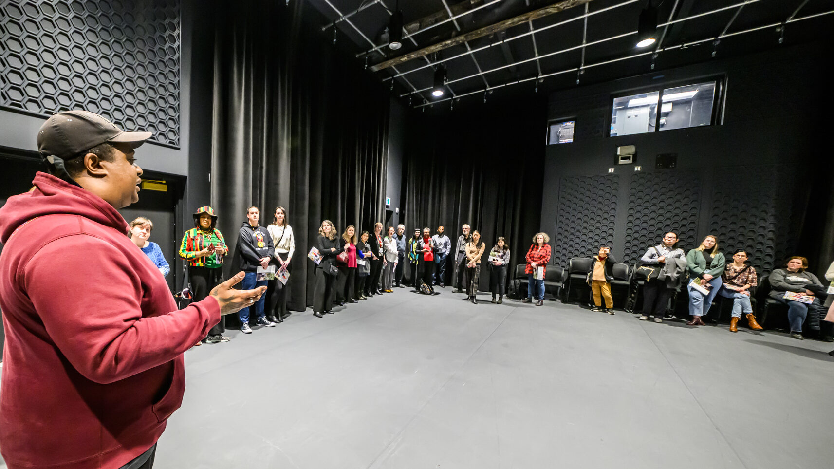 Person in foreground to the left addresses a group lined along two perpendicular walls in the background. Black curtain can be seen on the walls and an extensive lighting/equipment grid is partially visible on the ceiling.