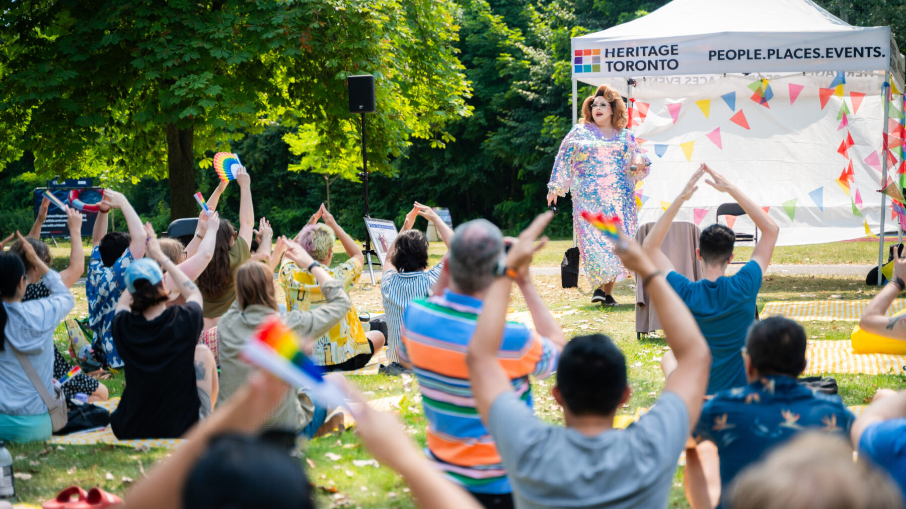 A drag queen in a shimmering and reflective sequins dress stands in front of a tent with the Heritage Toronto Logo emblem on it. A crowd of individuals seated on the grass before her raise their arms over their heads to make a tent shape under the instruction of the drag queen.