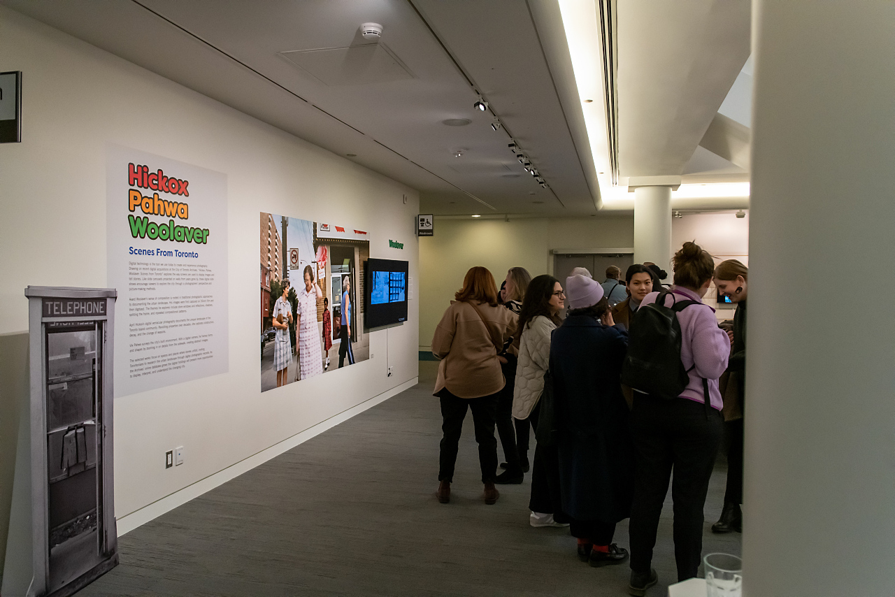 An art exhibit. The left side of the image shows an art installation on the wall. Text on the wall reads "Hickox Pahwa Woolaver: Scenes from Toronto" and is followed by smaller text. Beside the text is a large photograph and a small TV screen. The right side of the image shows a group of people talking and looking at the art.