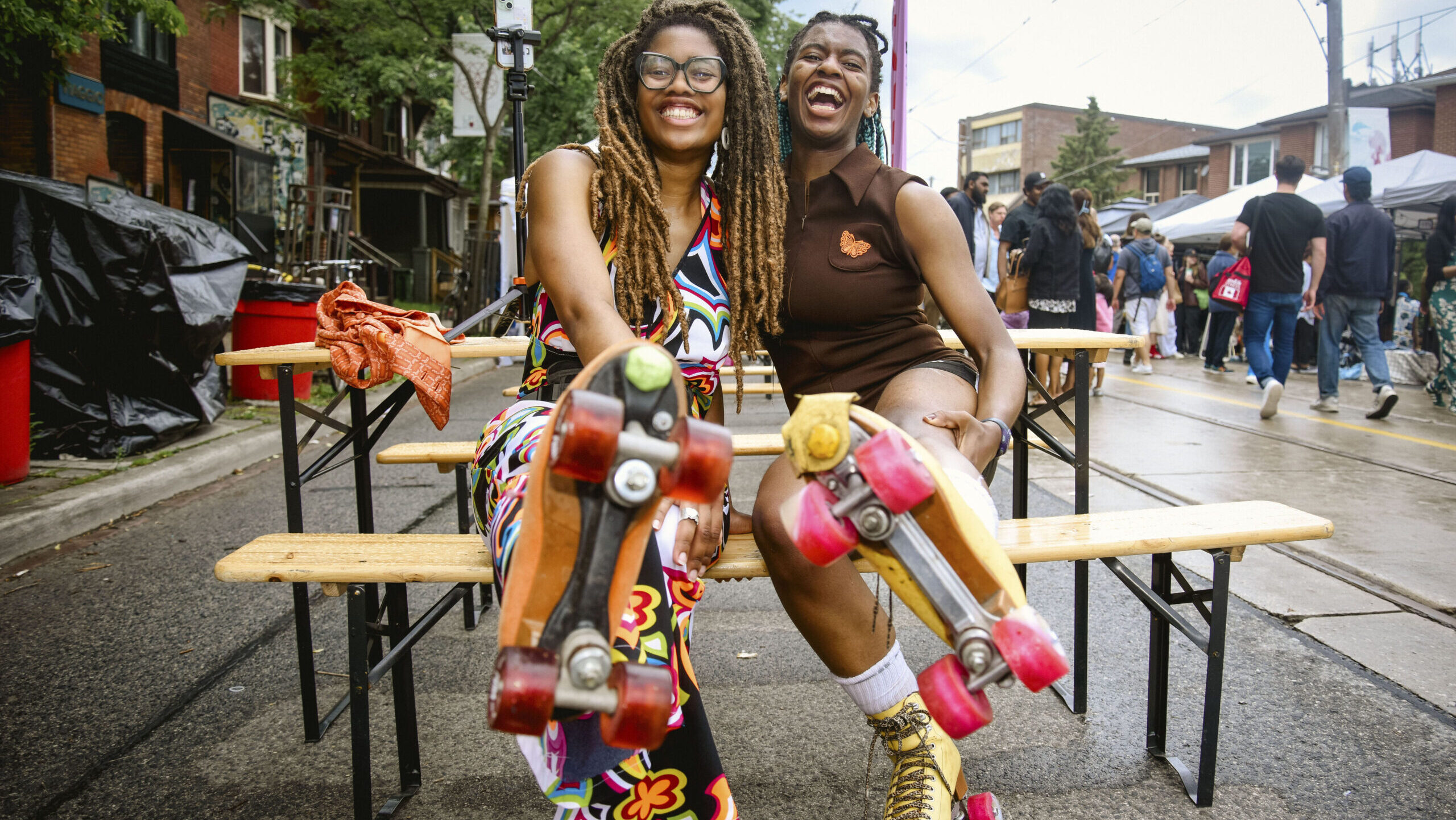 Two smiling Black women sitting on a bench, hugging with one leg raised to show their roller skates. The bottom of the roller skates are the focal point.
