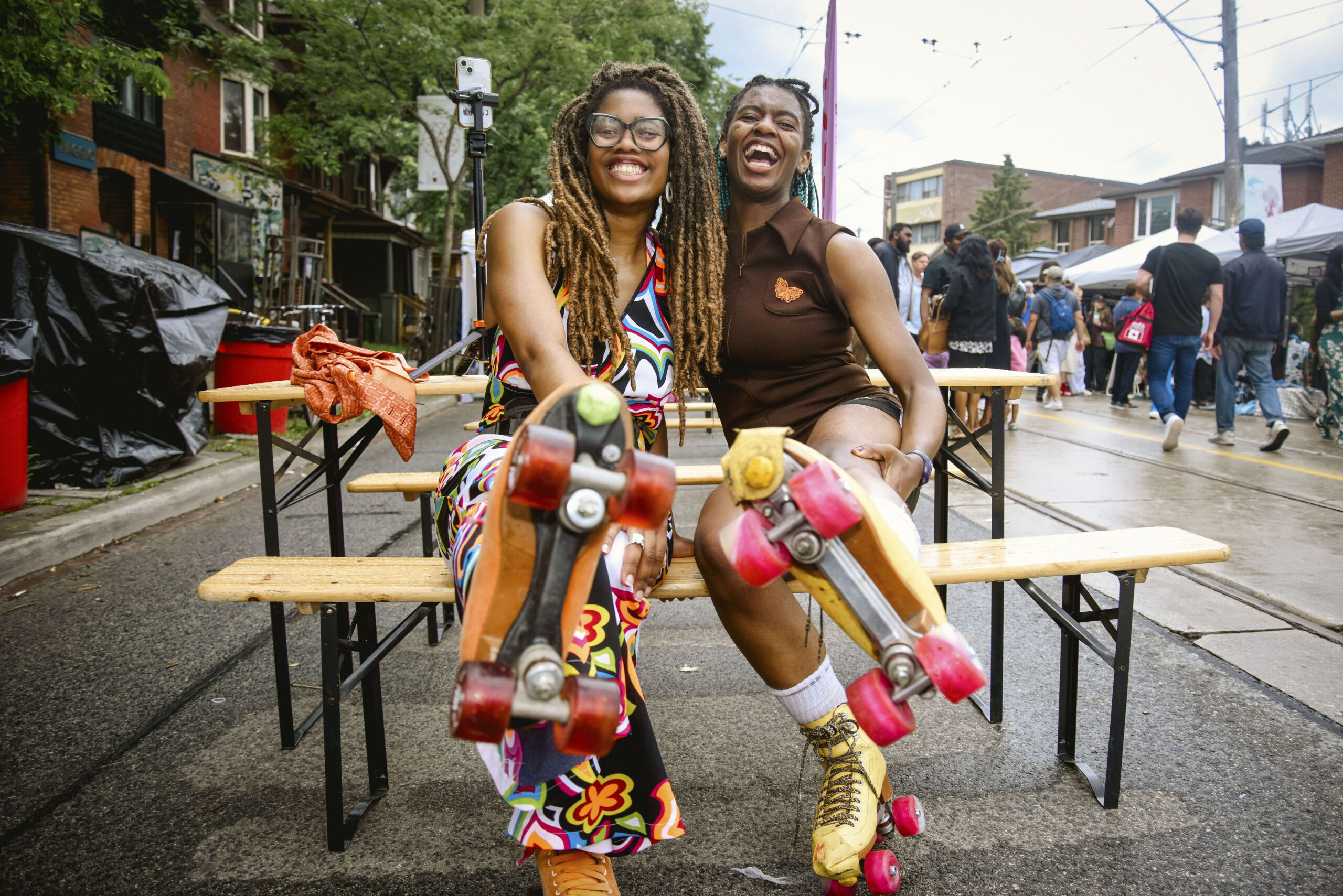 Two smiling Black women sitting on a bench, hugging with one leg raised to show their roller skates. The bottom of the roller skates are the focal point.