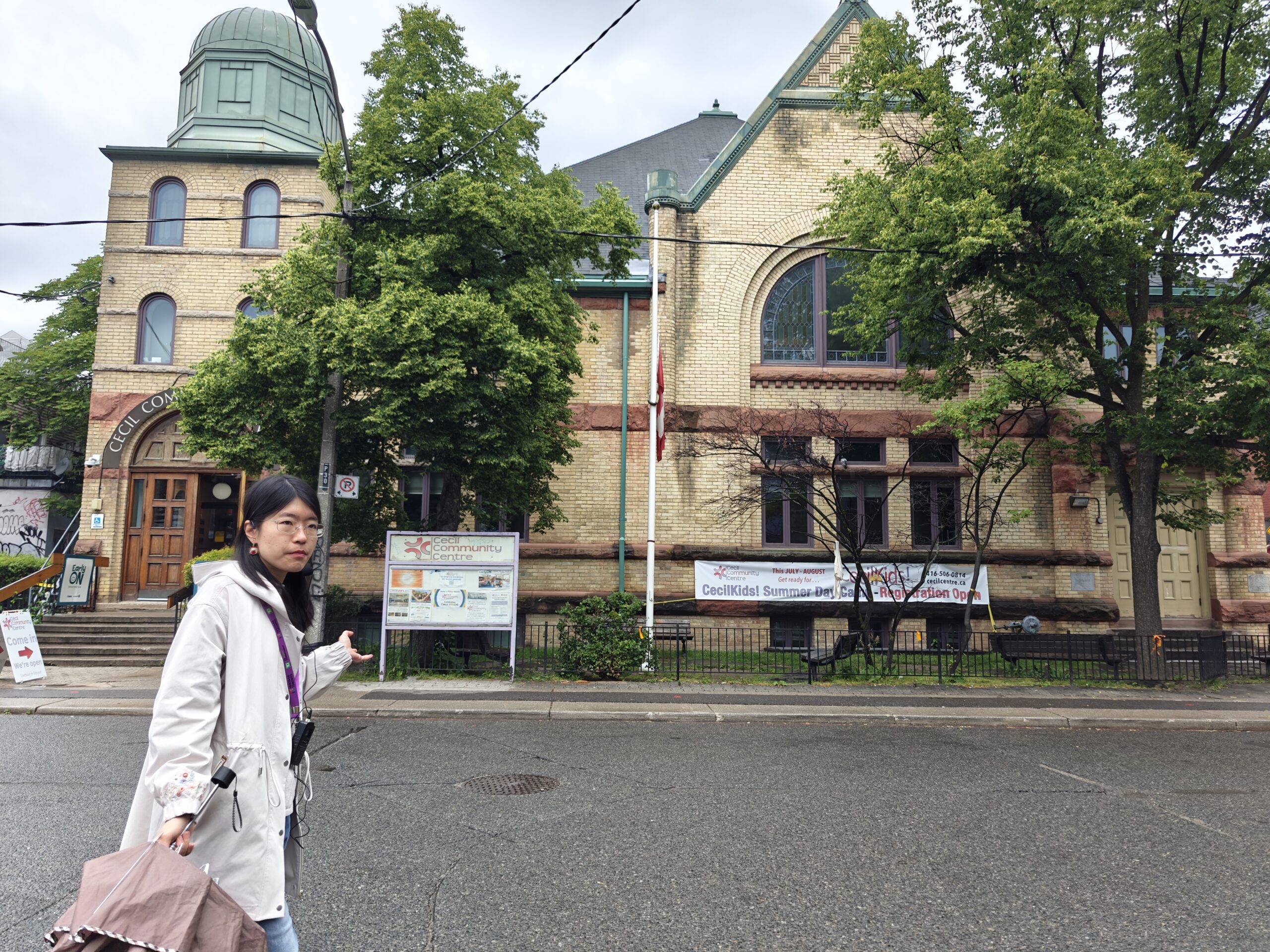A young woman walks across frame in front of a large brick building with a yellow tinge and green roof (similar to the green colour of oxidizing metal) across the street from her. In front of the building are two deciduous trees and a flag pole with the Canadian flag that is not raised.