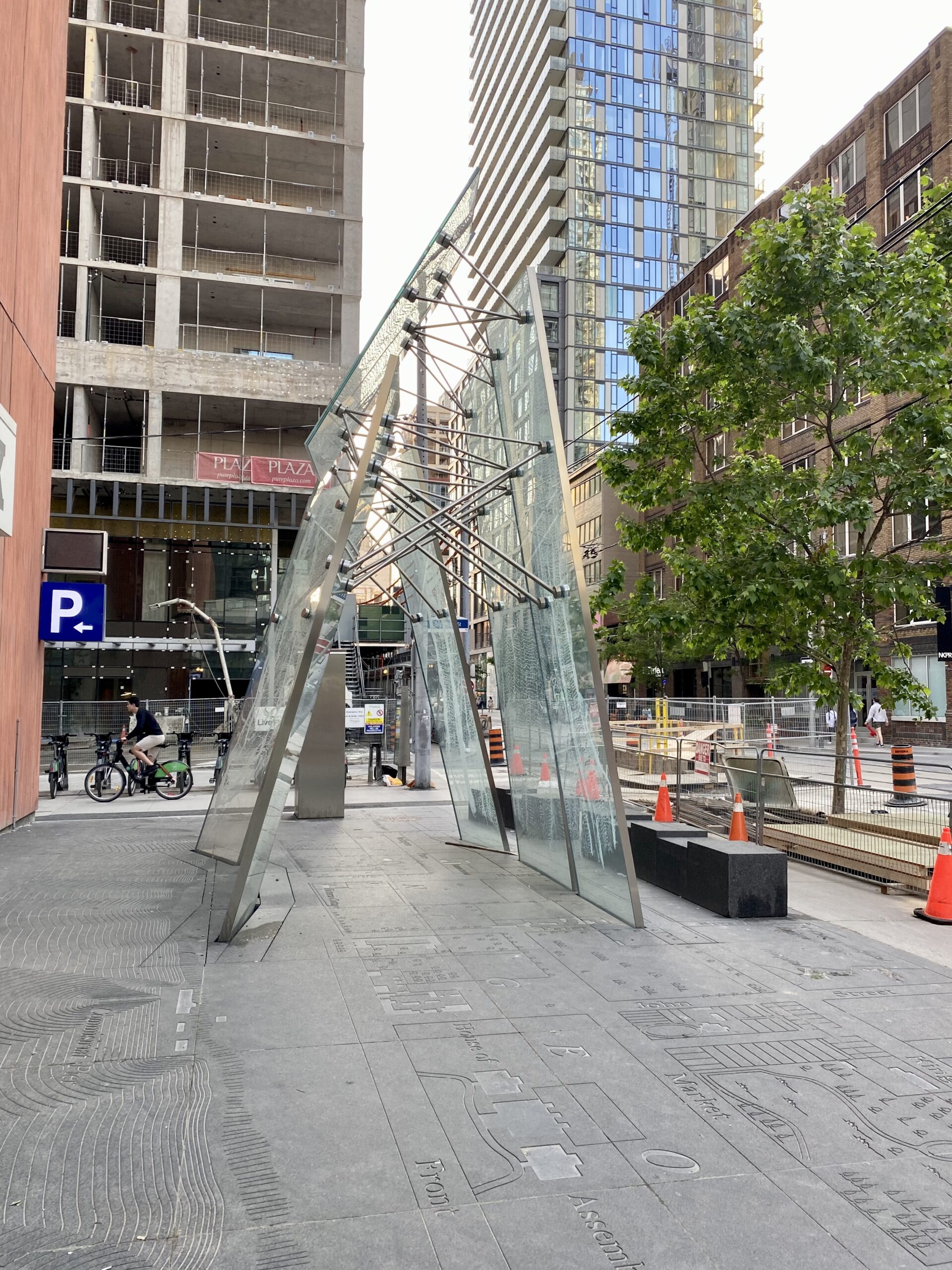Image of a tent-like structure made of glass and metal. Below is a stone surface that has been engraved with a map of part of downtown. To the right of the structure are some black stone benches.