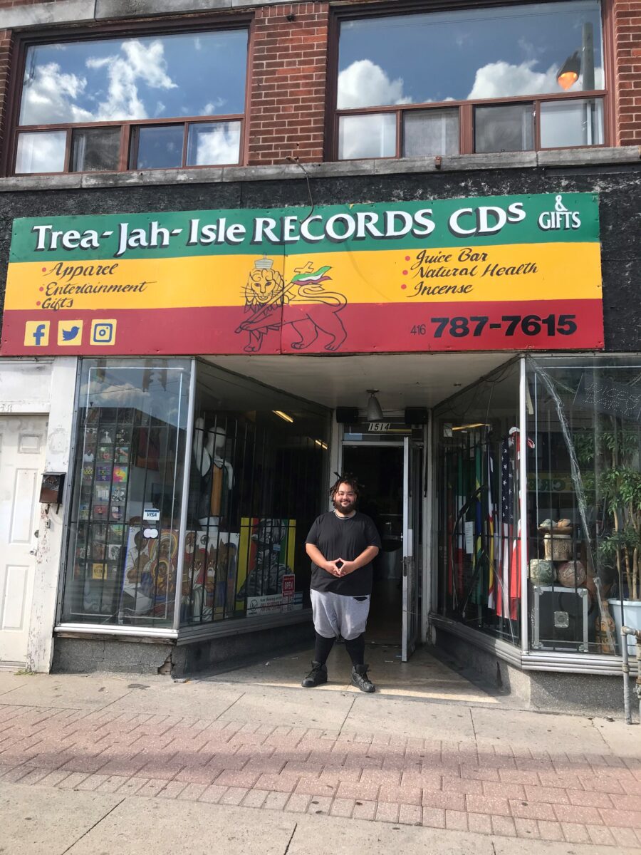 Photo of a man standing in front of a shop with his hands together in front of him smiling at the camera. Various items are visible in the windows of the shop on either side of the doorway, and the shops overhead green, yellow, and red sign reads, "Trea-Jah-Isle Records CDs". The sign also reads, "Apparel/ Entertainment/ Gifts/ Juice Bar/ Natural Health/ Incense/ 416-787-7615". In the centre of the sign is the image of a lion with a crown on carrying a cross over its shoulder. The sign is green, yellow, and red.