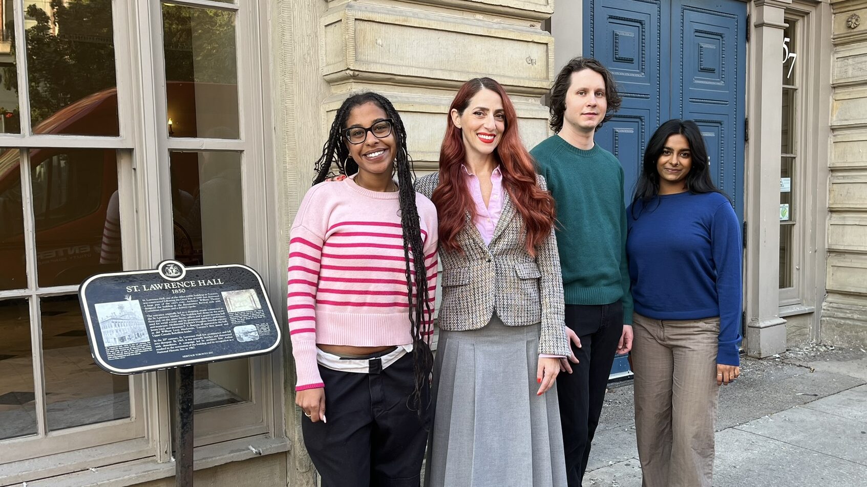 Four people stand outside a building entrance with blue doors.  On the left, a plaque mounted on a post is visible.